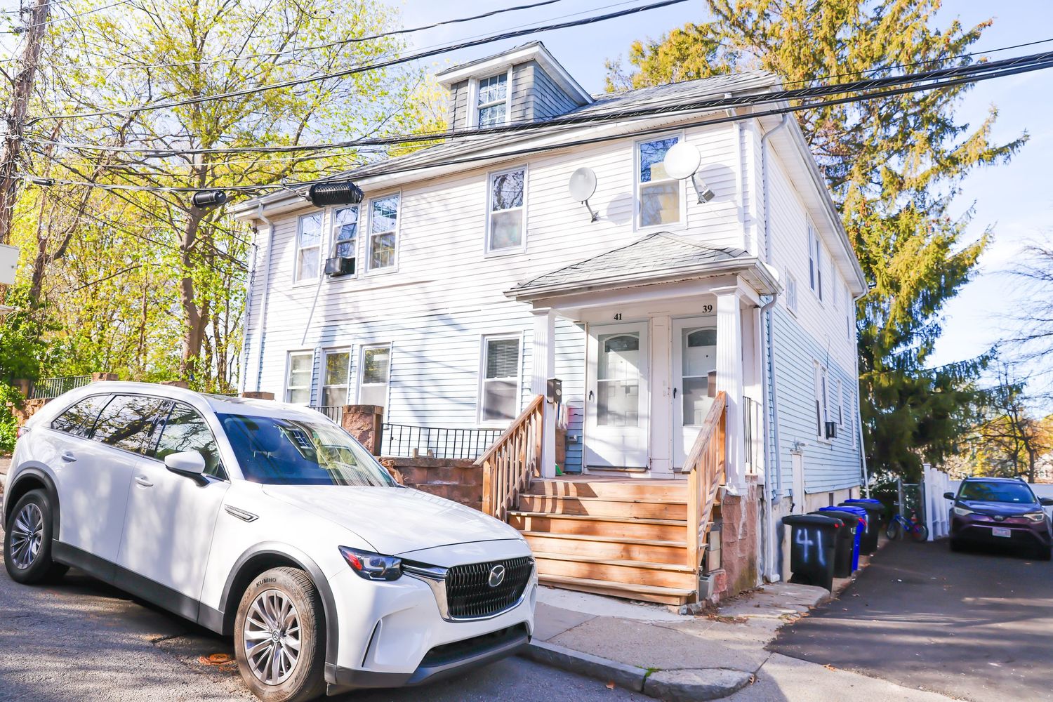 White two-story colonial home with front porch steps, white Mazda SUV parked in driveway, surrounded by spring trees.