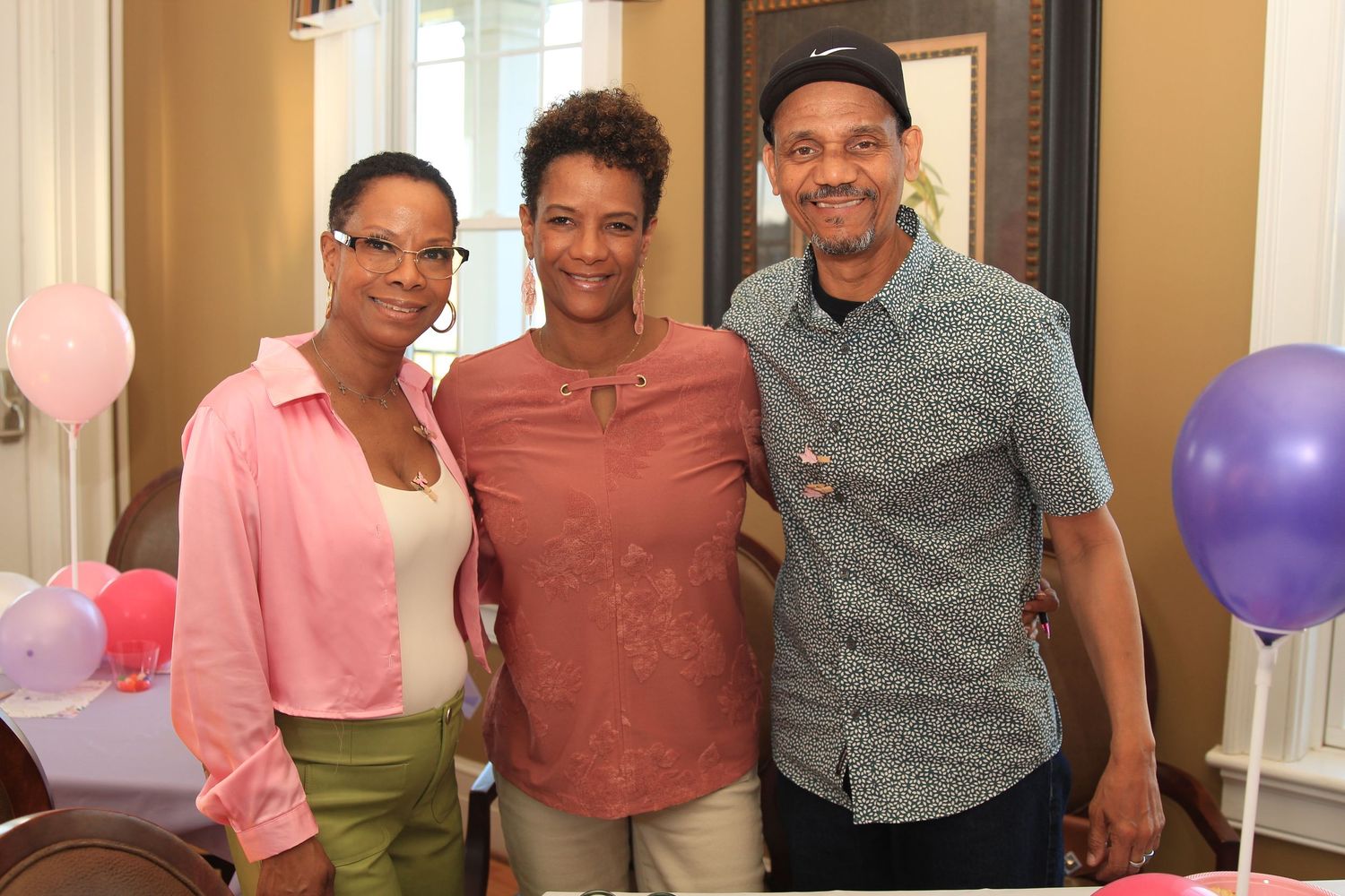 Three adults smiling together at a pink and purple balloon-decorated baby shower event.