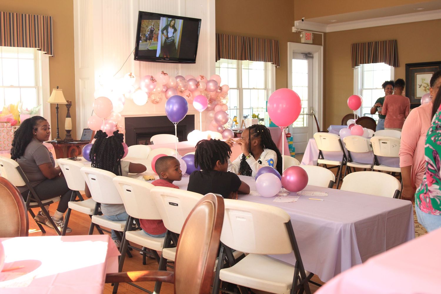 Guests sit at balloon-decorated tables during a pink and purple themed birthday celebration in a banquet hall.