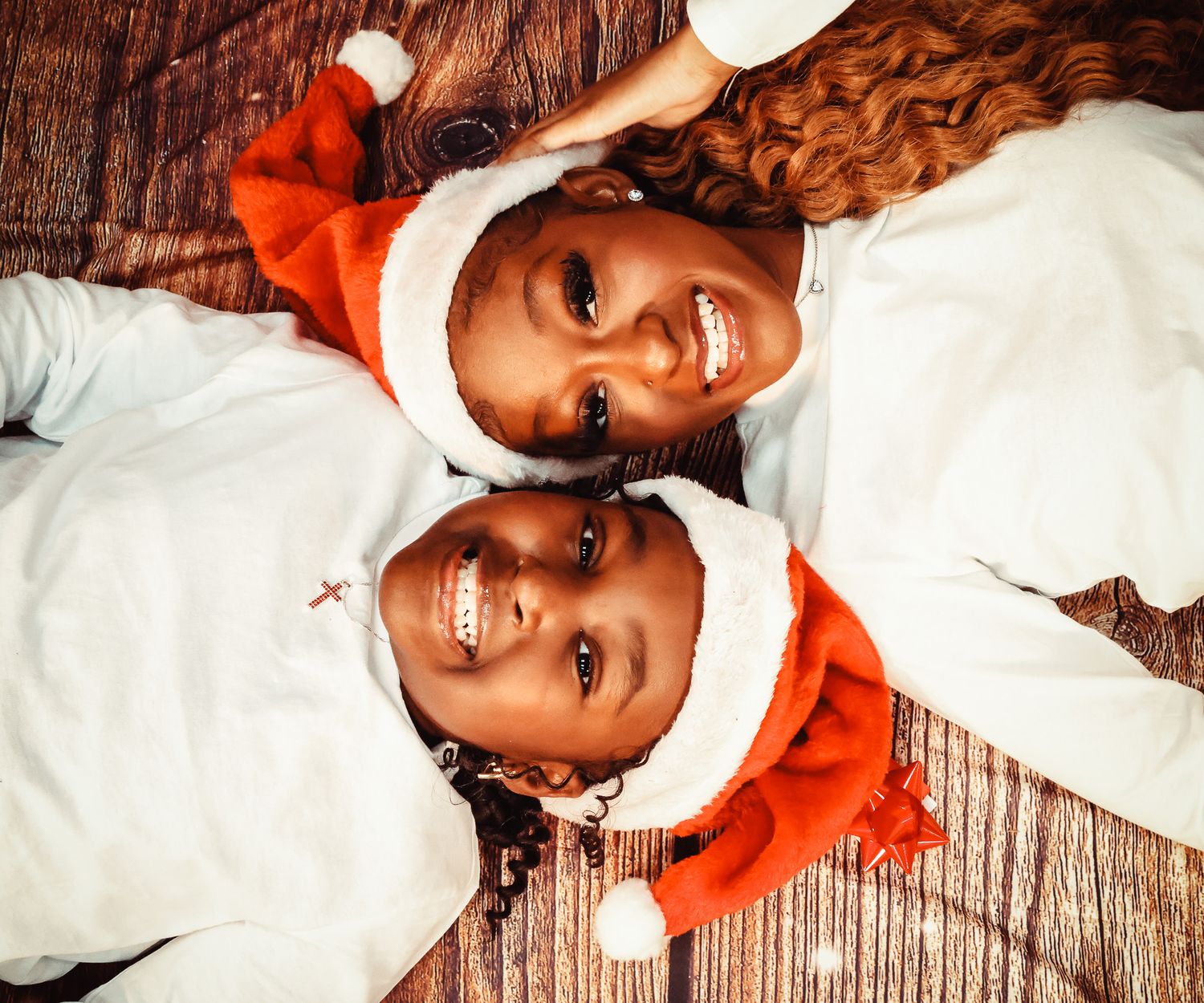 Two children wearing Santa hats and white outfits smiling while lying on wooden floor for Christmas photo.