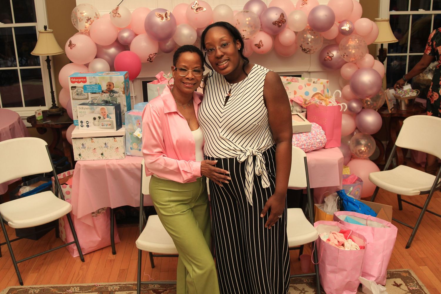 Two women embrace and smile at an elegant pink baby shower party with balloon arch backdrop.