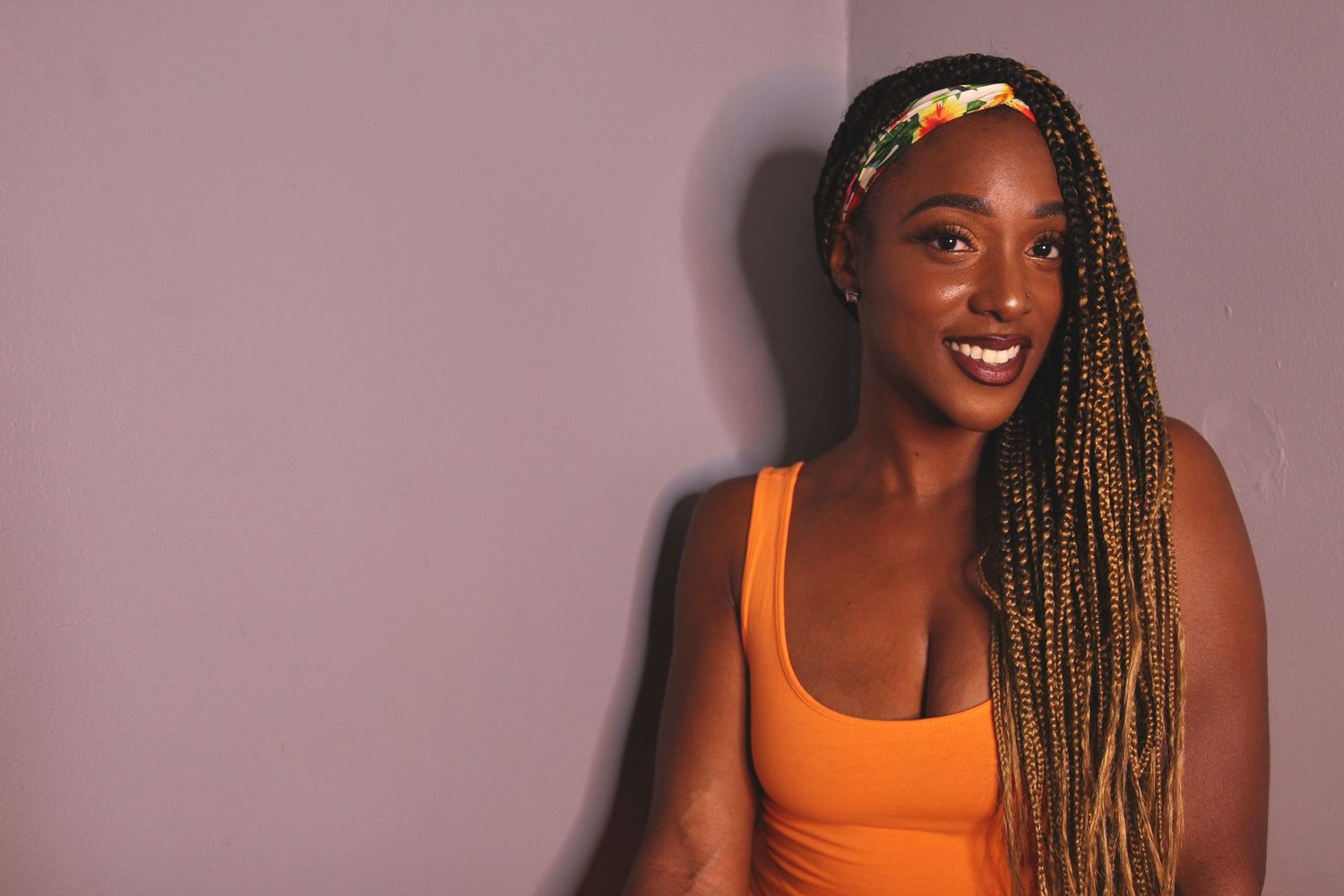 Smiling Black woman with long box braids and colorful headband wearing an orange tank top against a purple wall.
