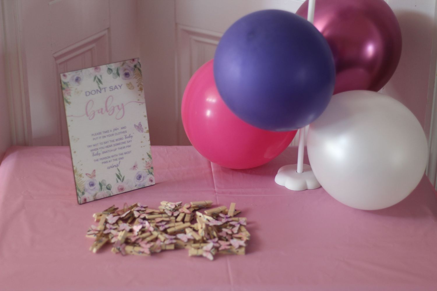 Pink table with purple, pink, and white balloons, a floral sign, and scattered wooden letter tiles for a baby shower.