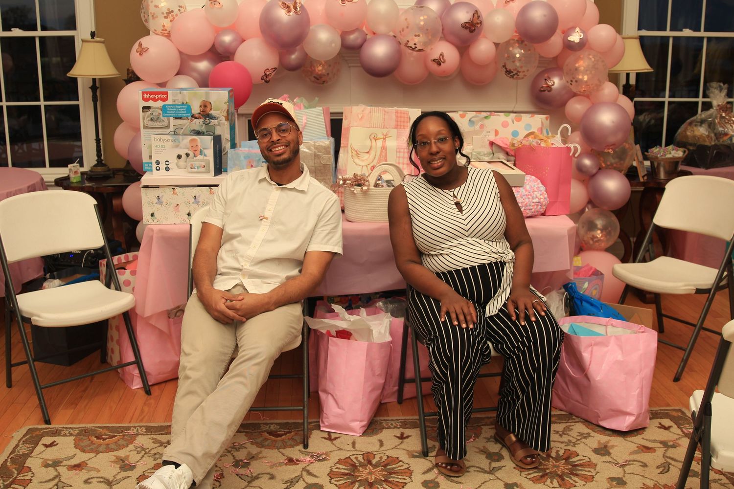 Couple posing together at a pink baby shower surrounded by gifts and balloon arch.