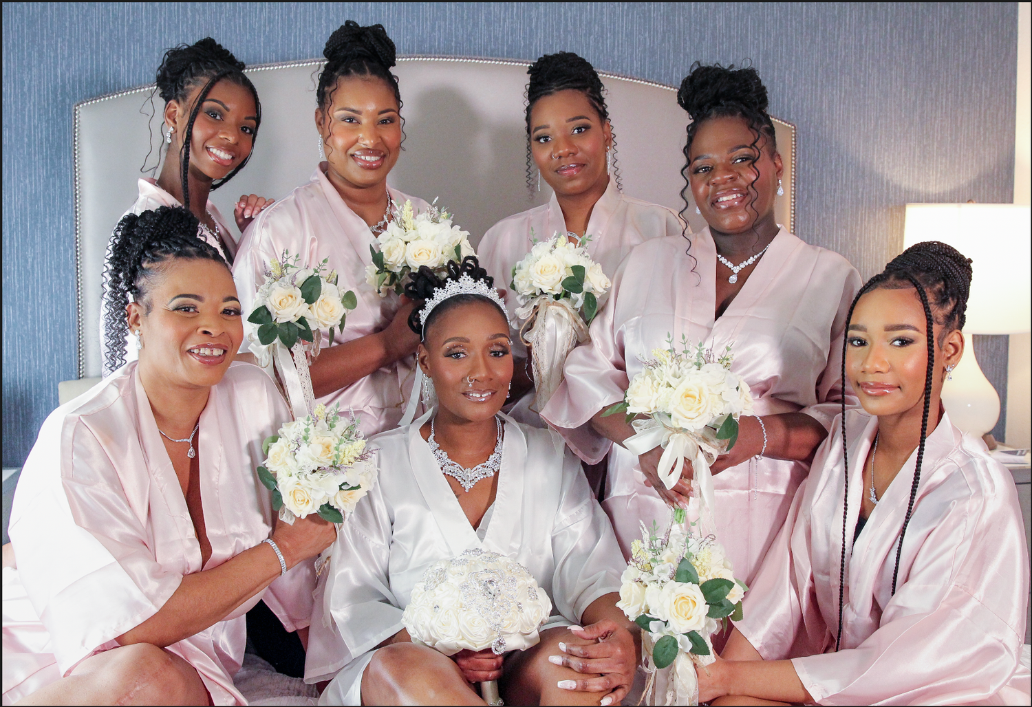 Bride with bridesmaids in pink robes holding white flower bouquets posing together on a bed before a wedding.