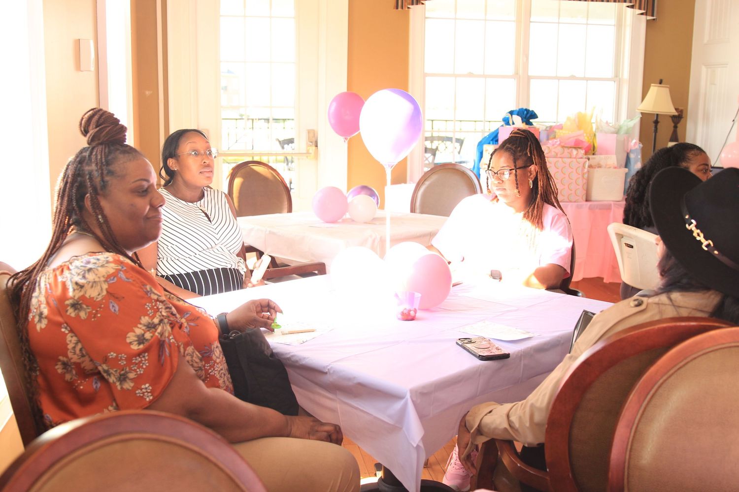Women chat at a white table adorned with pink and purple balloons during a birthday party celebration.