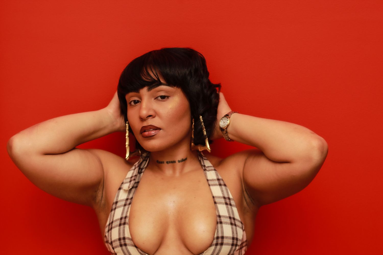 A woman in a plaid halter top poses with hands behind her head against a vibrant red background.