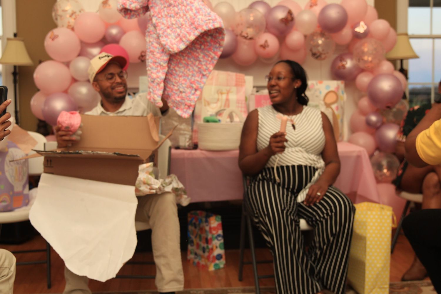 Couple opening gifts at a pink baby shower with balloon arch backdrop and wrapped presents.