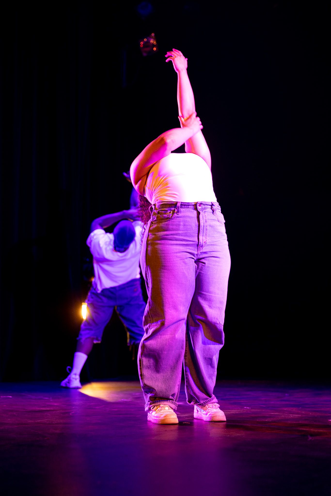 Dancer in white top and jeans performing on a colorfully lit stage with arms raised.