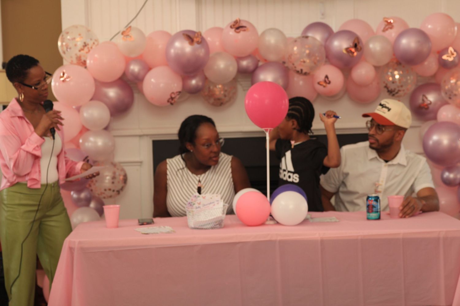 Group of people gathered around a pink table at a baby shower with balloon decorations.