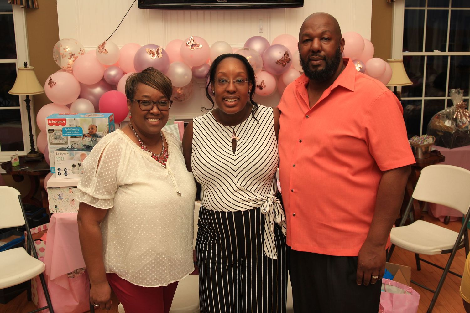 Three adults pose together smiling at a pink-themed baby shower party with balloon decorations.