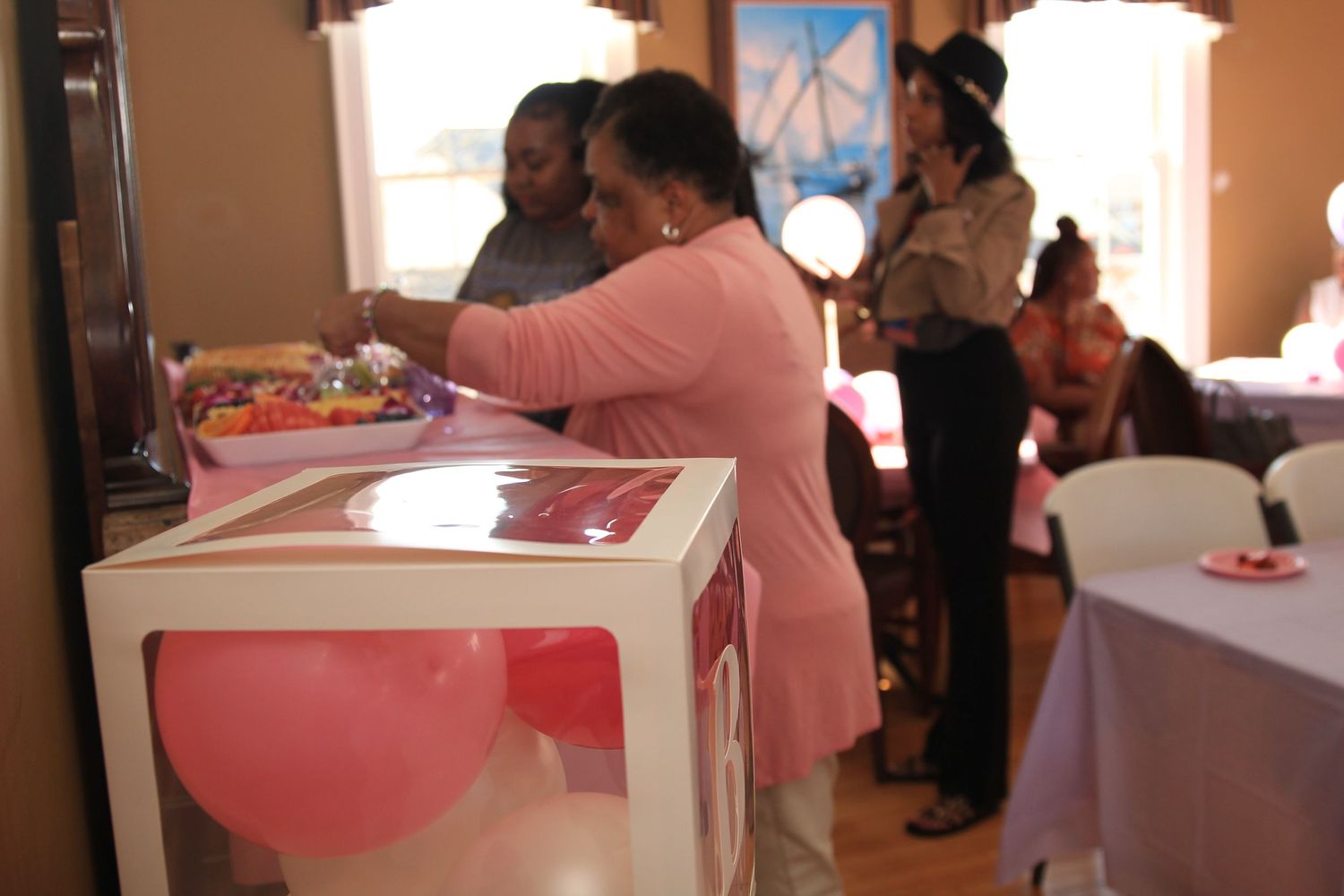 Woman in pink arranges decorations near a balloon-filled box at an indoor baby shower party.