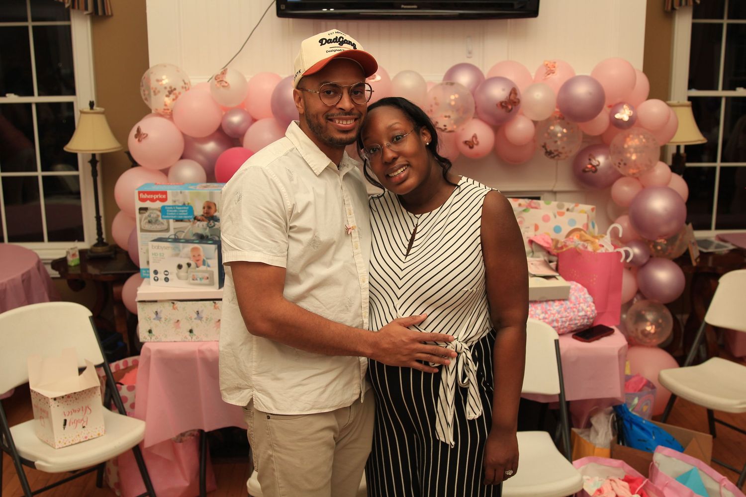 Couple embraces warmly at a pink baby shower with decorative balloon garland in background.