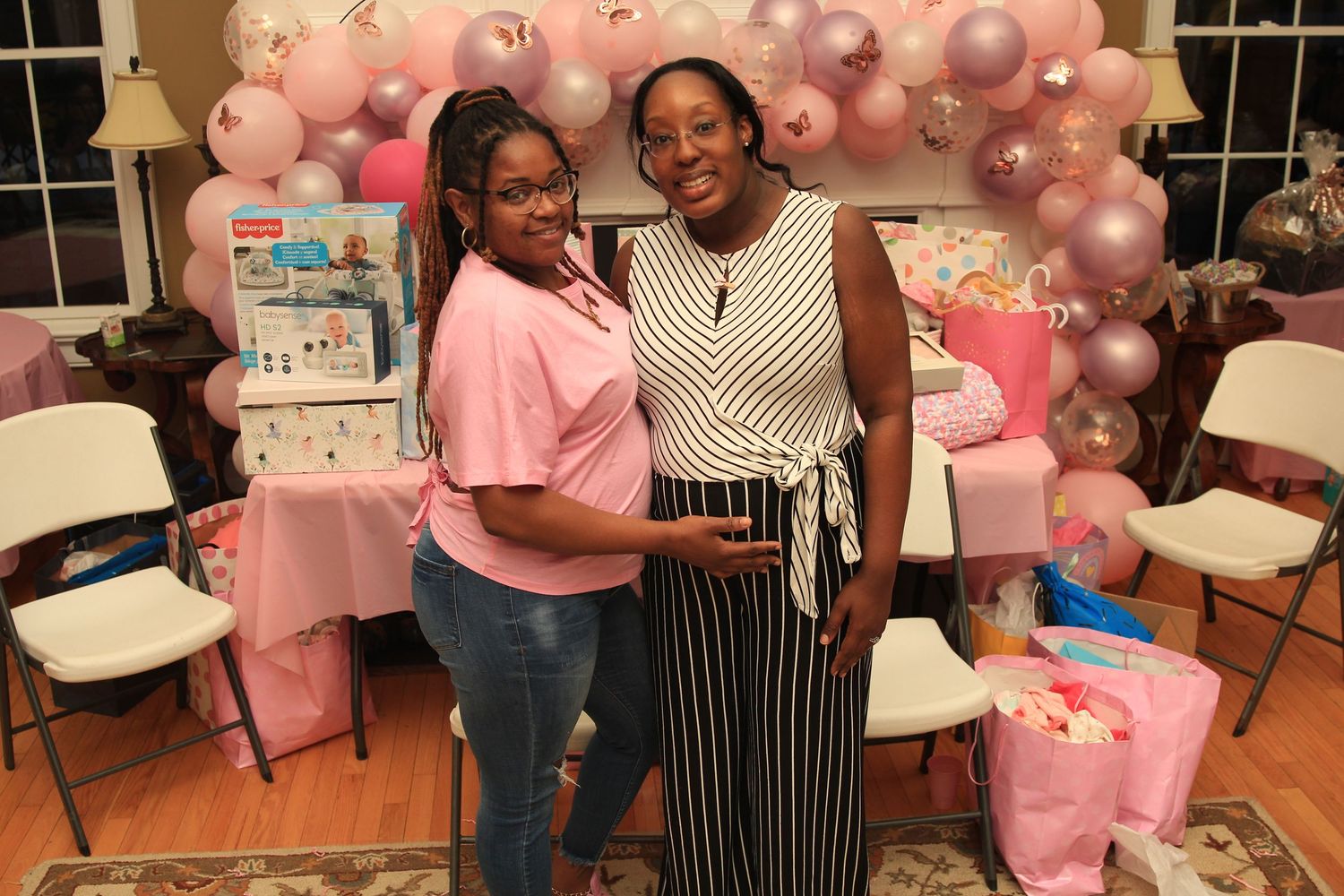 Two women pose together smiling at a pink-themed baby shower with balloon garland decorations.