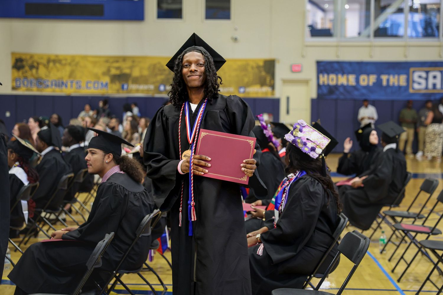 Graduate in black cap and gown proudly holds diploma at indoor graduation ceremony.