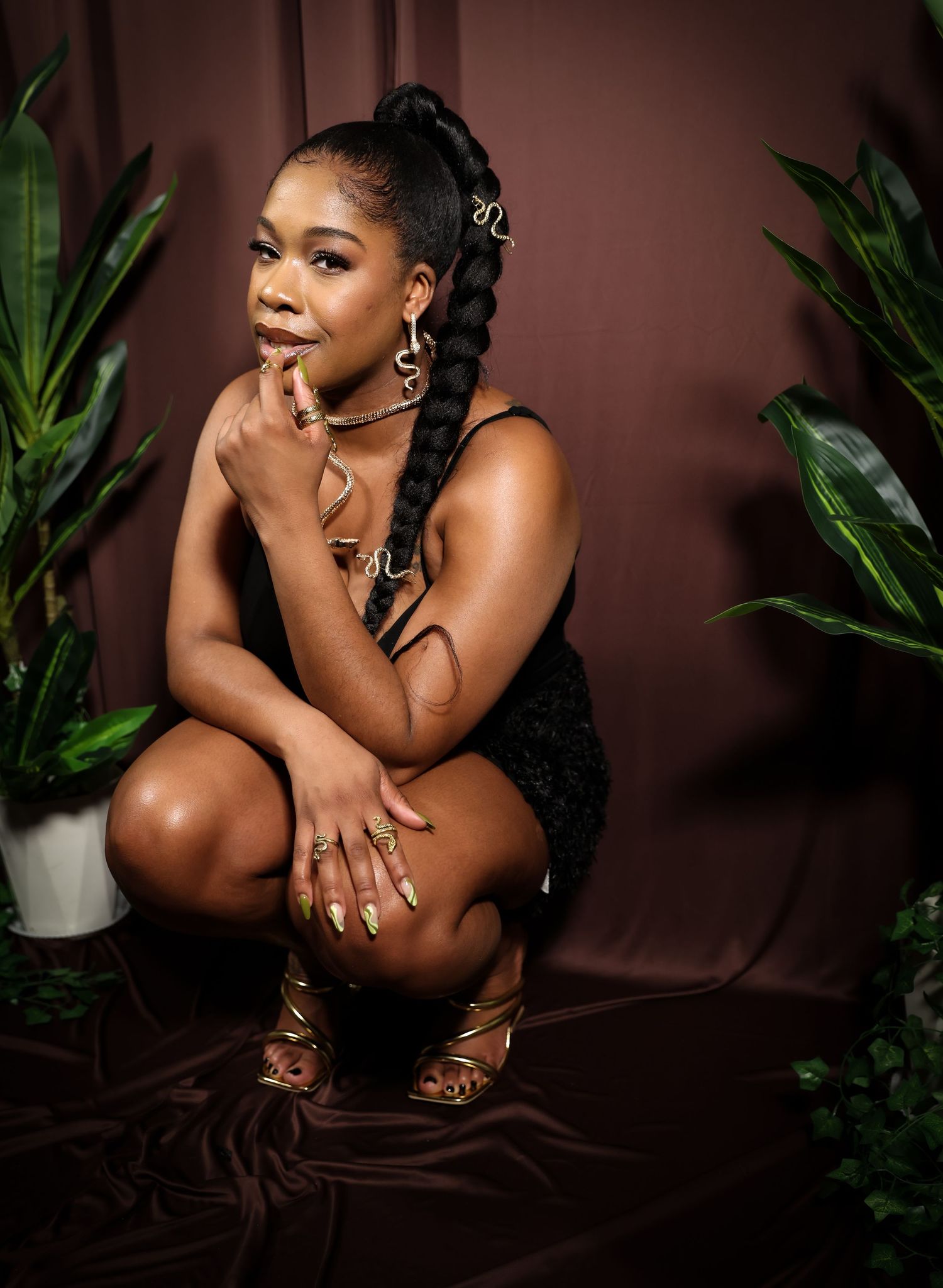 Woman with braided ponytail posing in a squat position wearing black outfit surrounded by green plants.