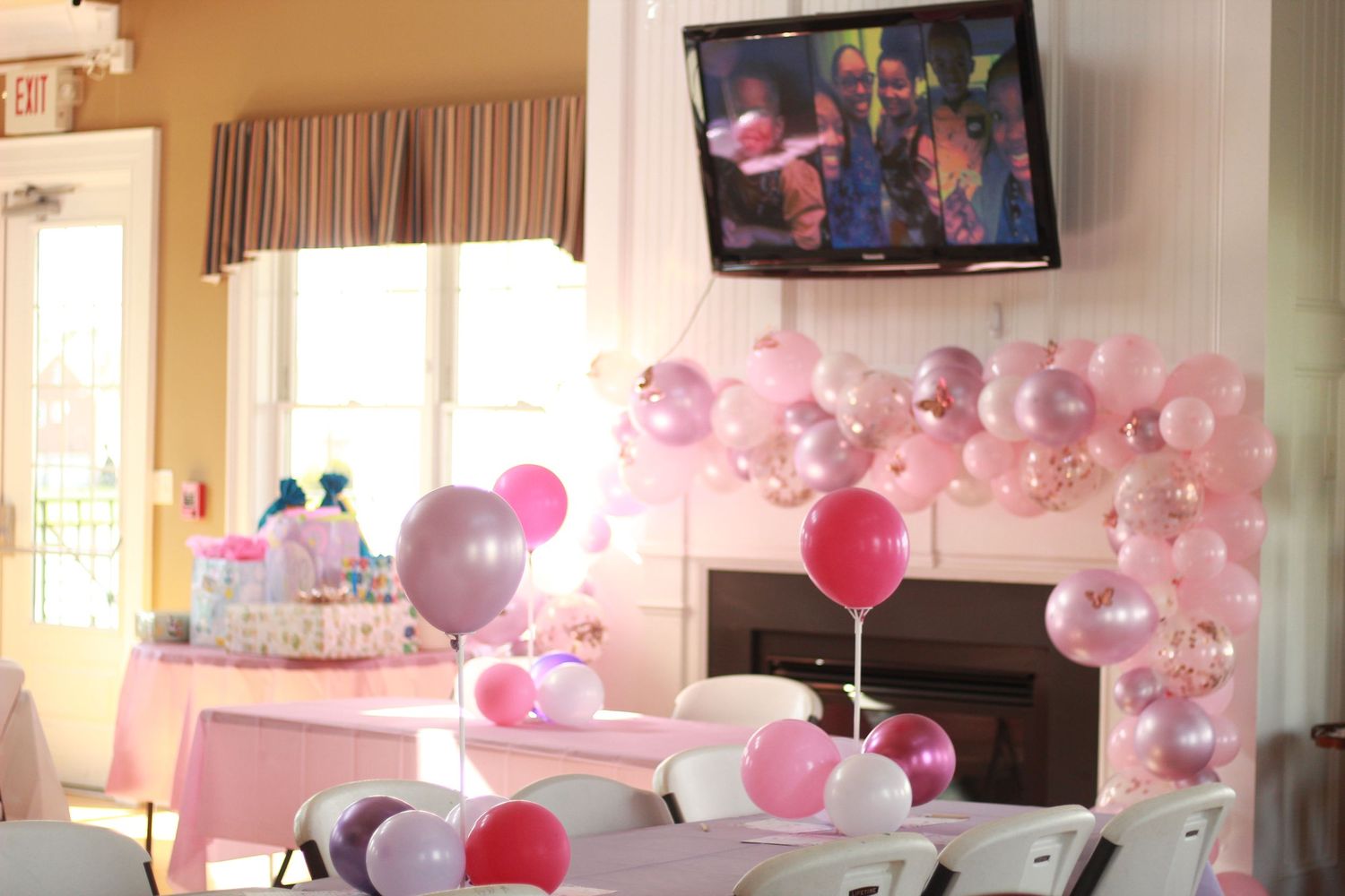 Pink and purple balloon arch decorating a baby shower venue with a TV mounted above a fireplace.