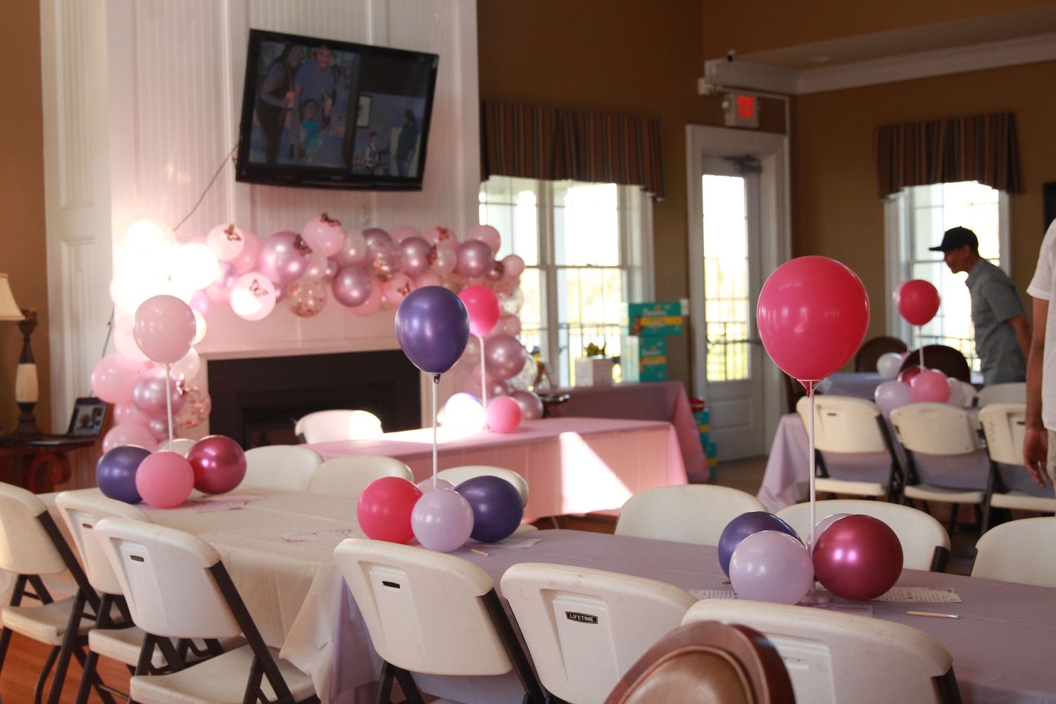 Festive pink and purple balloon centerpieces decorate white folding tables in a party venue with a TV screen.
