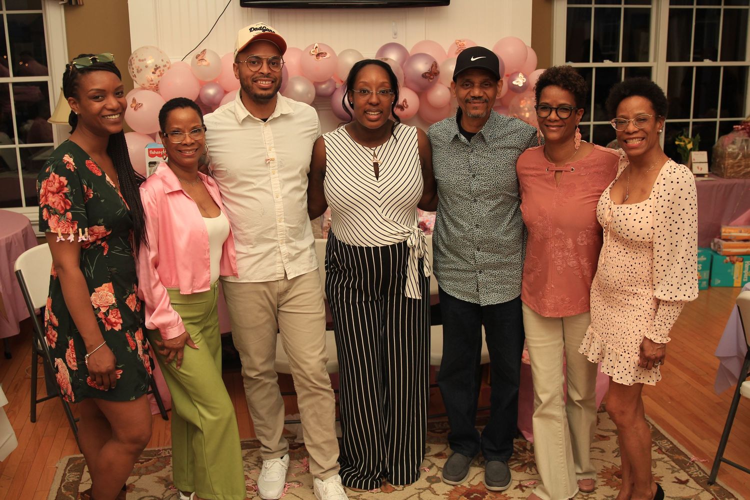 Group of seven people posing together at a festive pink baby shower with balloon garland backdrop.