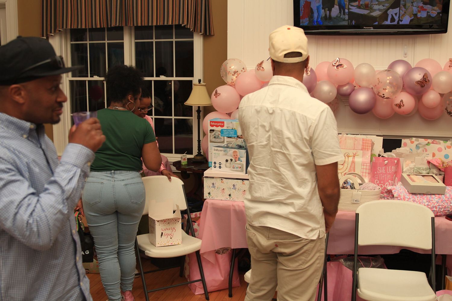 Guests browse baby shower gifts and decorations at a pink-themed celebration with balloon backdrop.