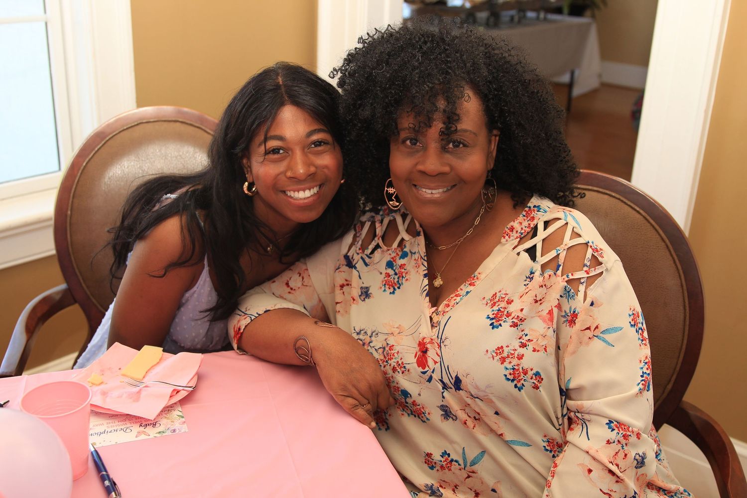 Two women smiling and posing together at a table during a pink-themed baby shower.
