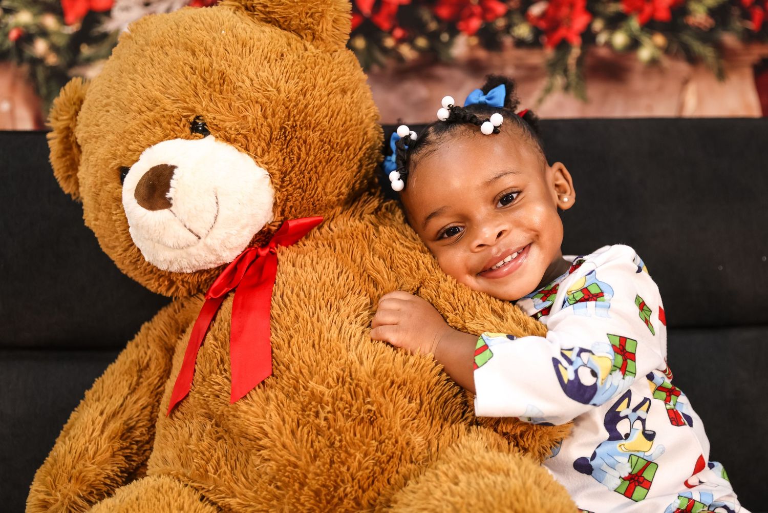 A smiling young girl hugs a large brown teddy bear with a red bow, wearing a festive outfit with blue hair bows.