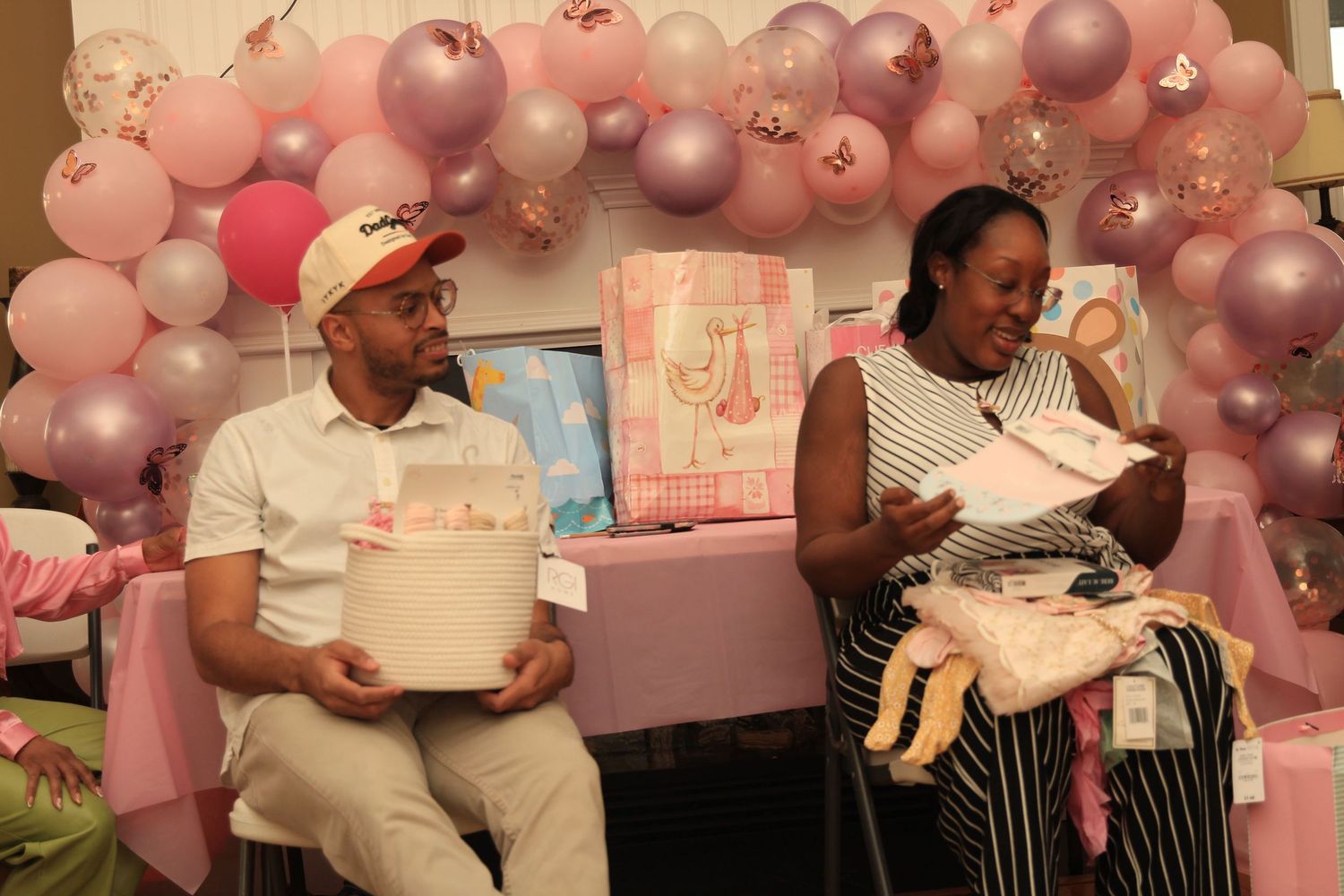 Couple opens baby shower gifts in front of a pink balloon arch.