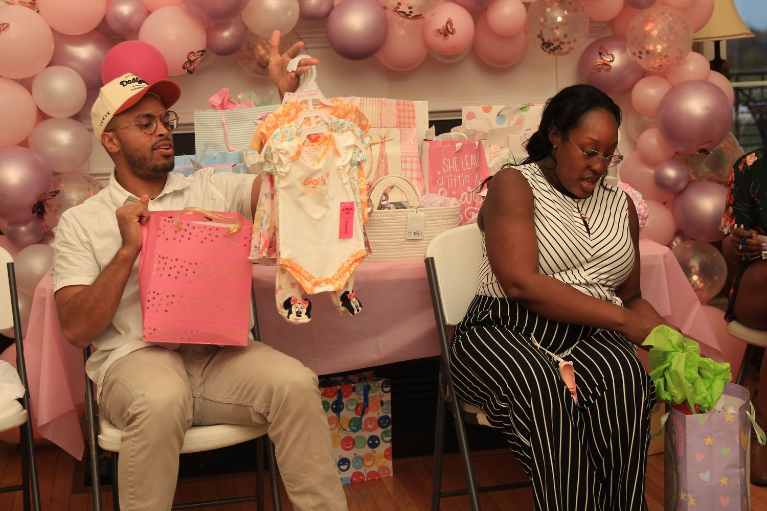 Man and pregnant woman opening pink gift bags at a decorated baby shower with balloon arch.