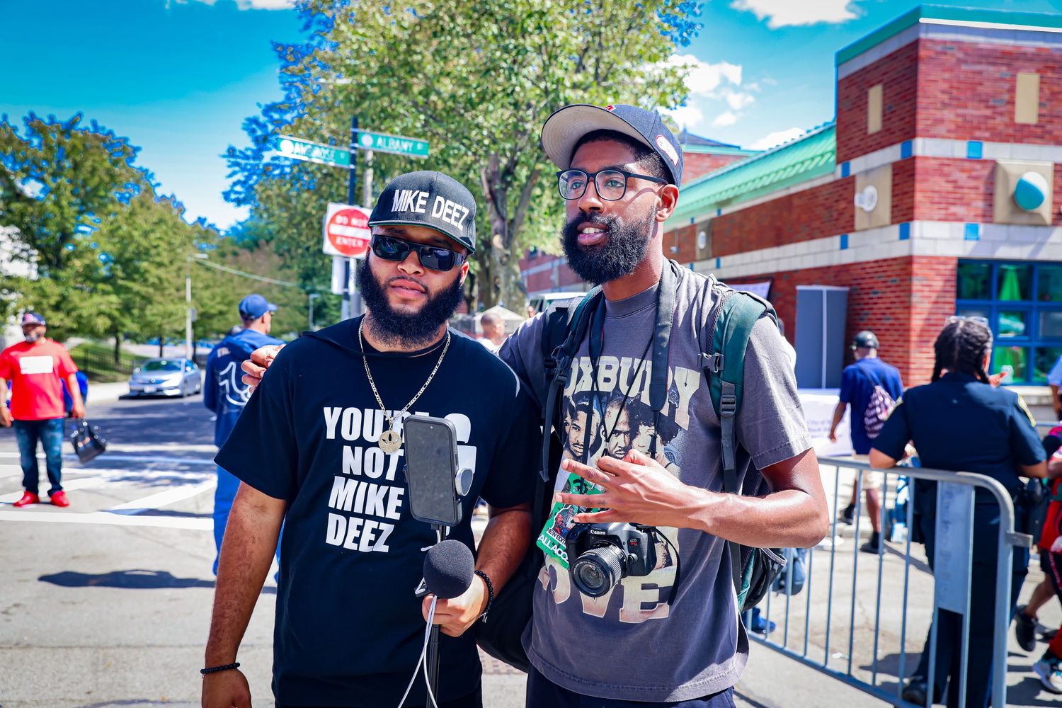 Two Black men posing outdoors at a street event, one wearing a Mike Deez t-shirt and the other holding a mic.