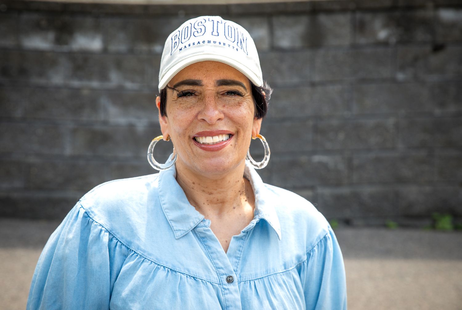 A smiling woman in a light blue shirt and white trucker cap stands outdoors near a stone wall.