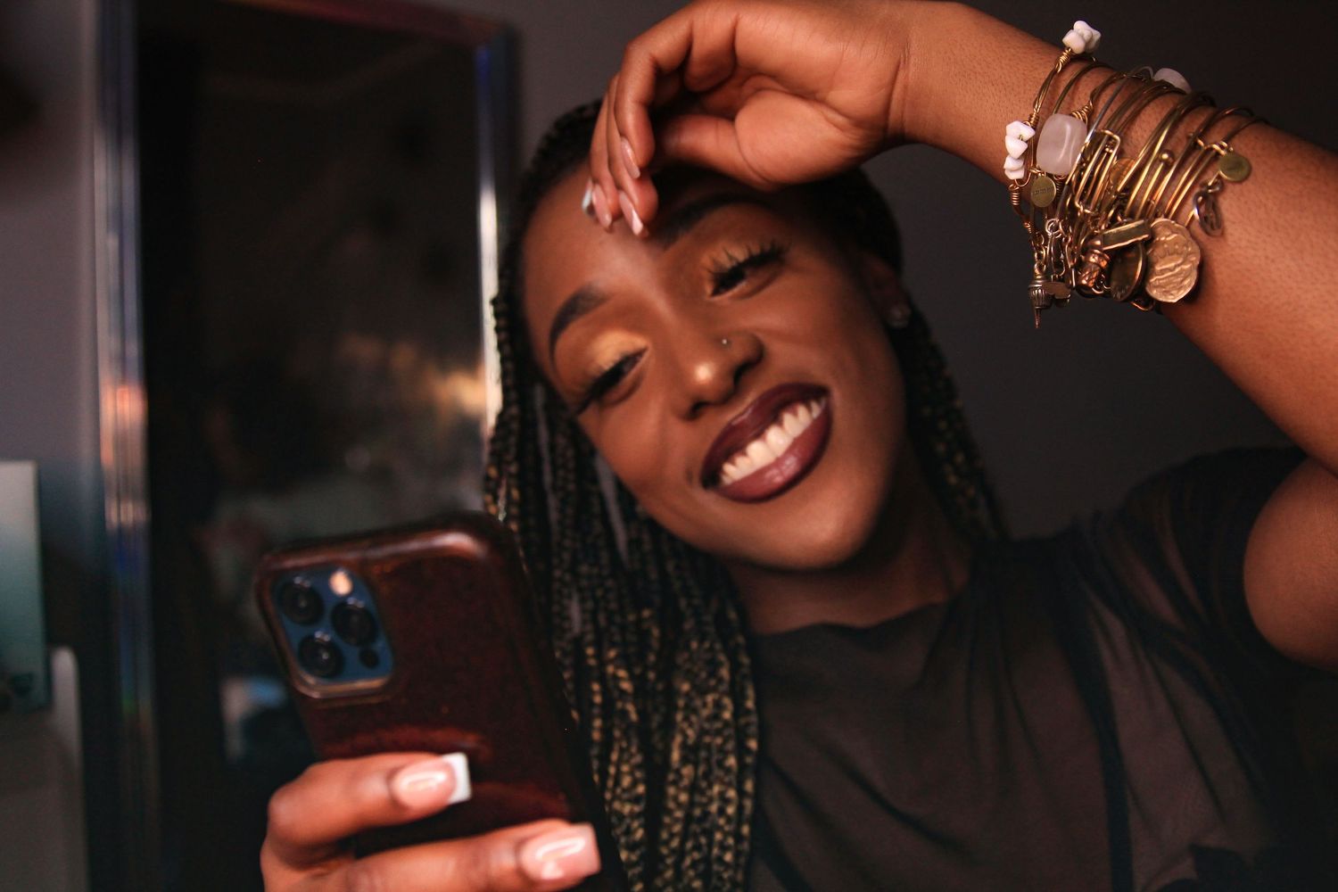 Smiling young Black woman with braids taking a selfie, wearing gold bangles, holding a smartphone.