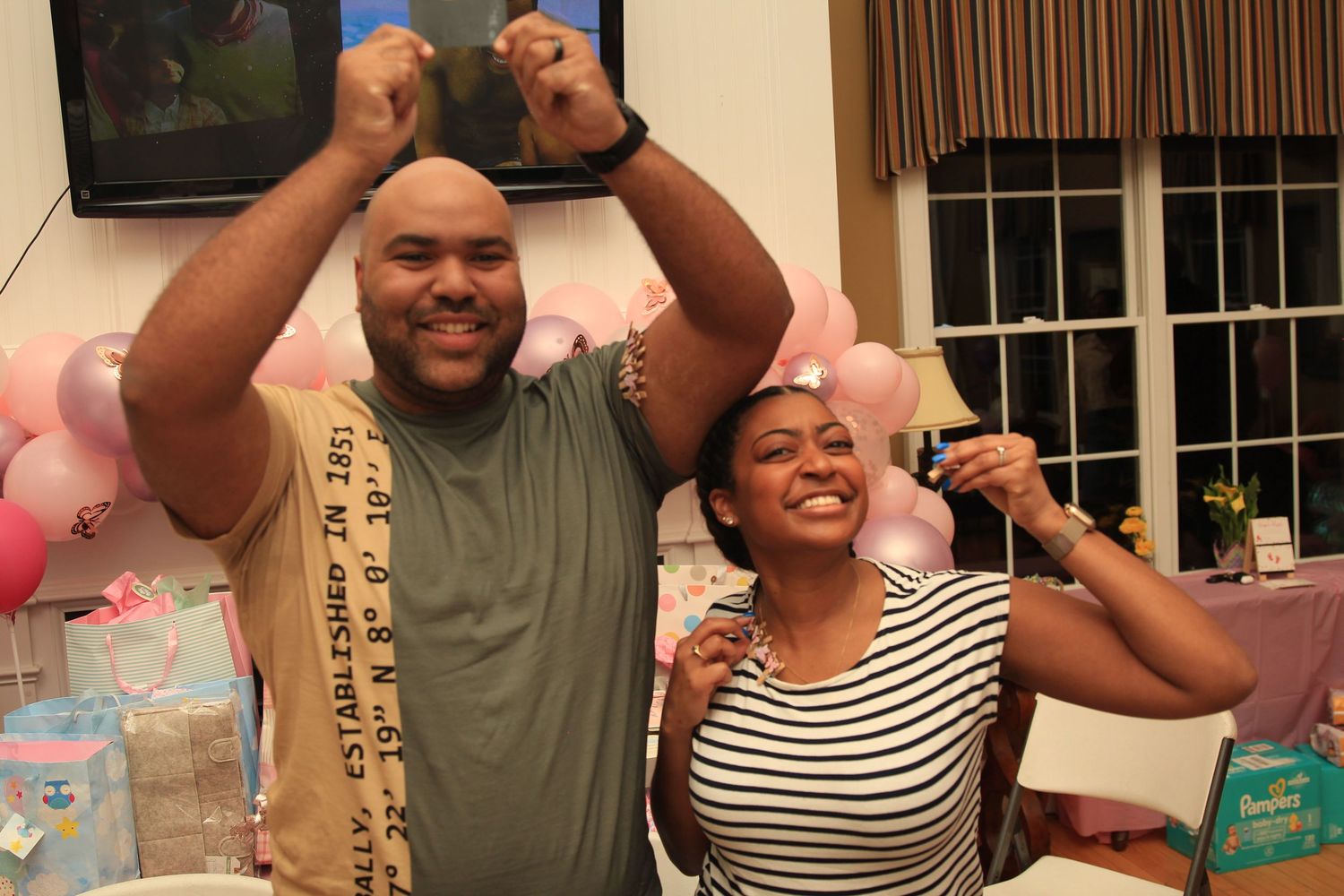 Excited couple celebrating at a pink-themed baby shower with balloons and decorations in the background.