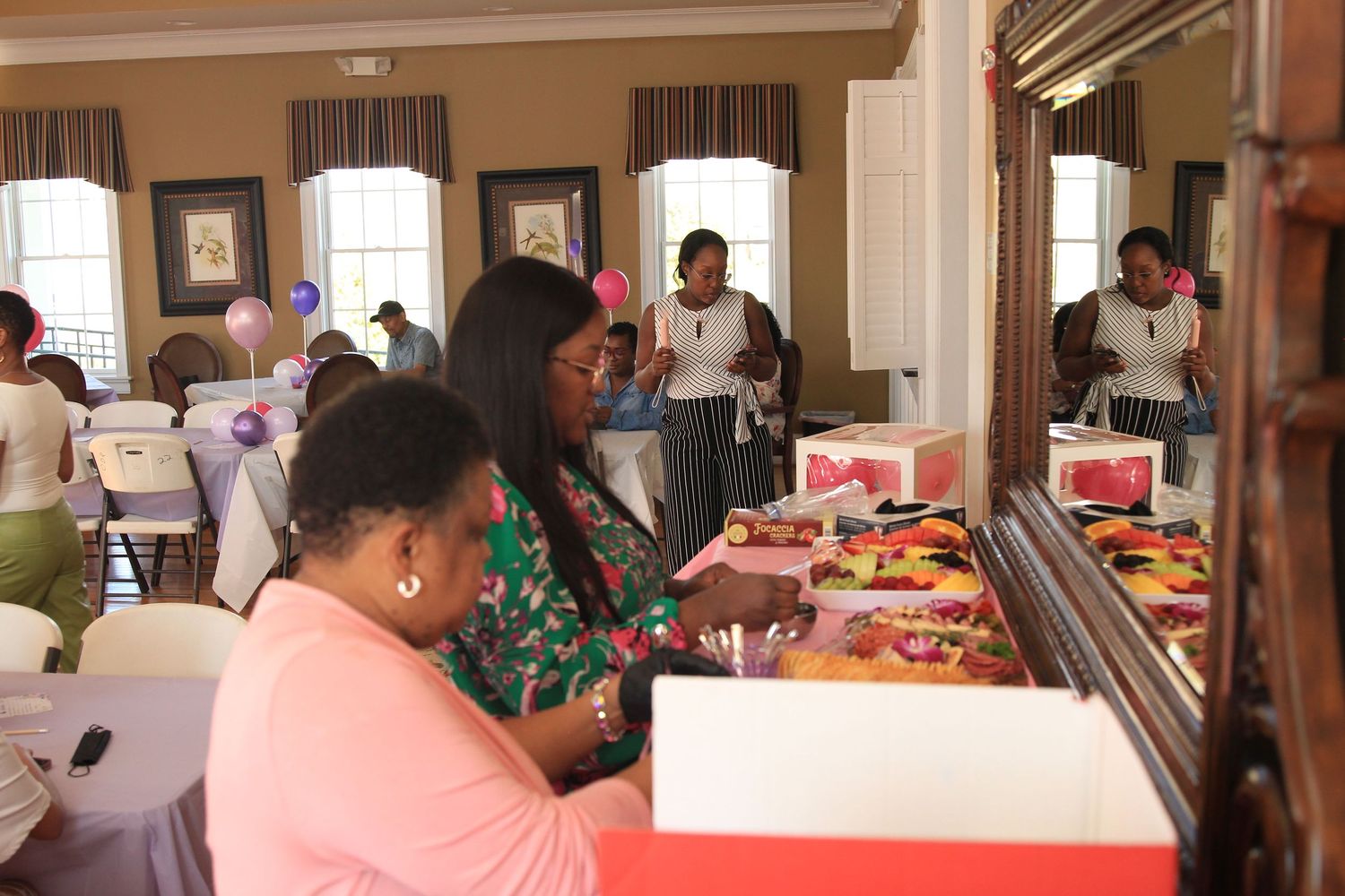 Two women examine items on a decorated table at a pink-themed birthday party in an elegant venue.