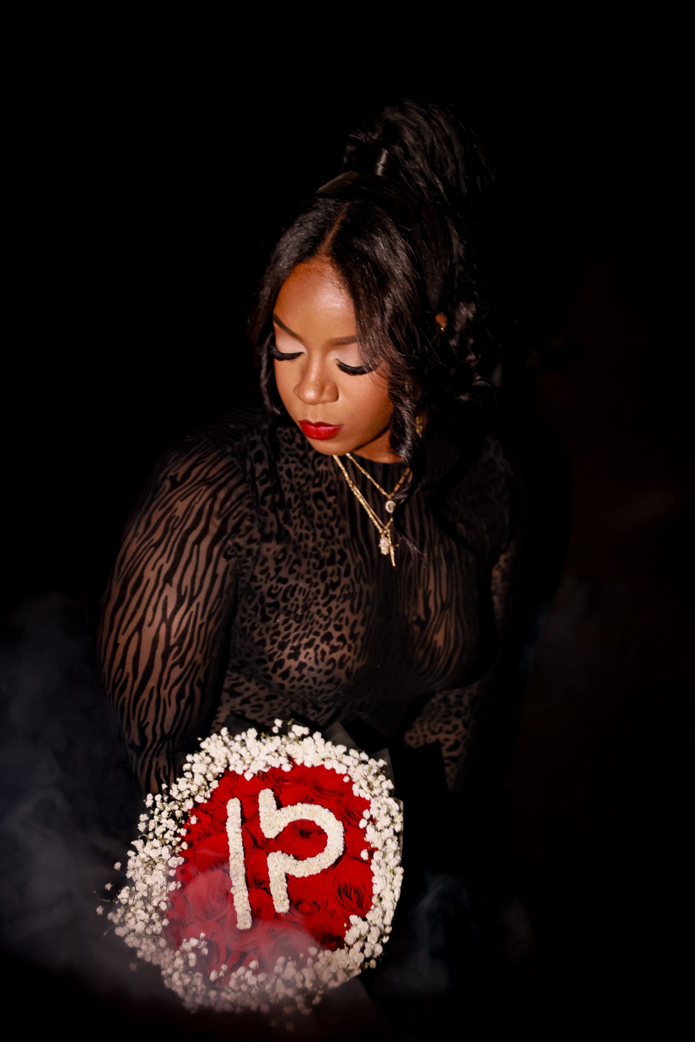 Woman in sheer lace dress holds glowing floral arrangement with number 15 in dramatic dark setting.