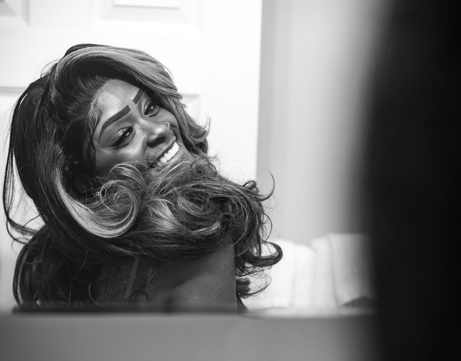 Black and white photo of a smiling Black woman tossing her long curly hair in front of a mirror.