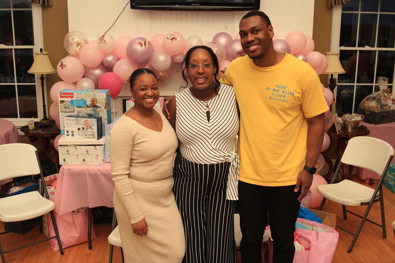 Three people smiling together at a baby shower with pink balloon arch and baby gifts.