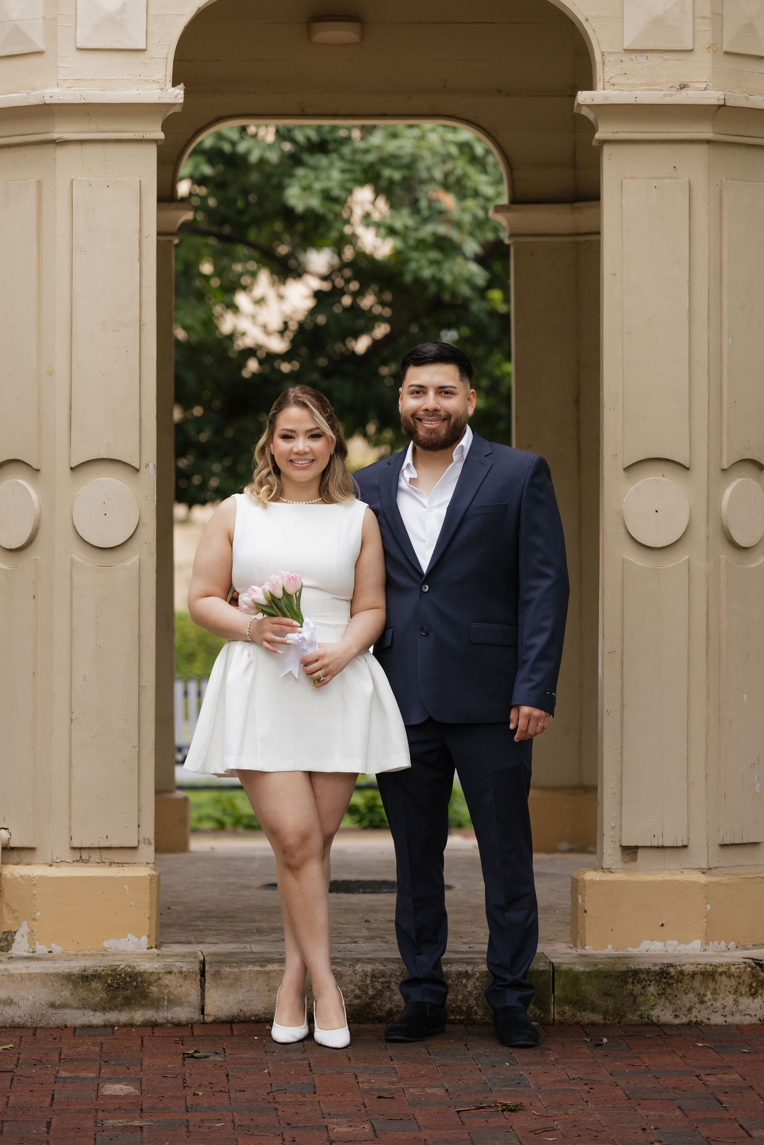 Couple in wedding attire posing under stone archway with small bouquet.