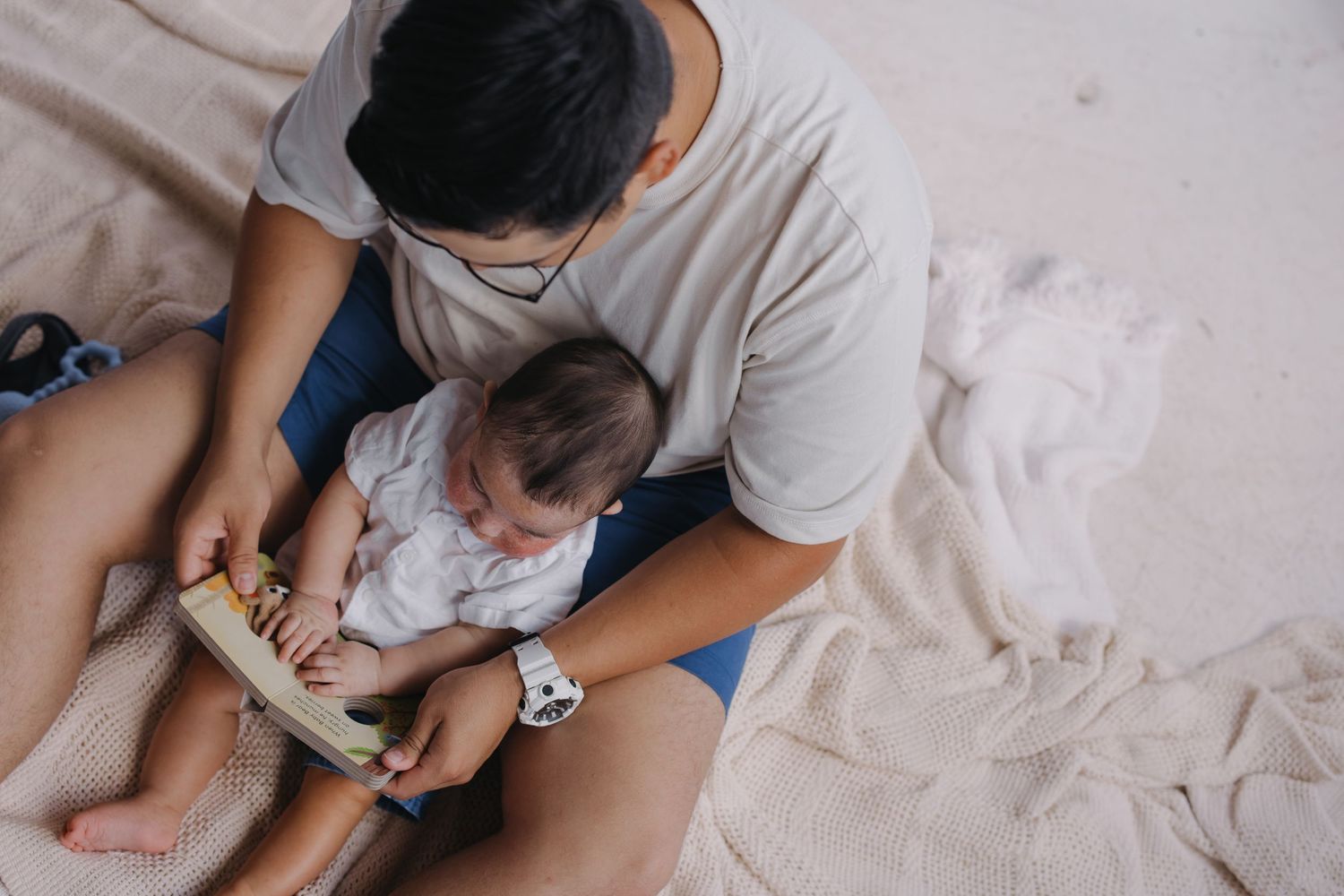 Father wearing glasses reading a book to newborn baby while sitting on a blanket indoors.