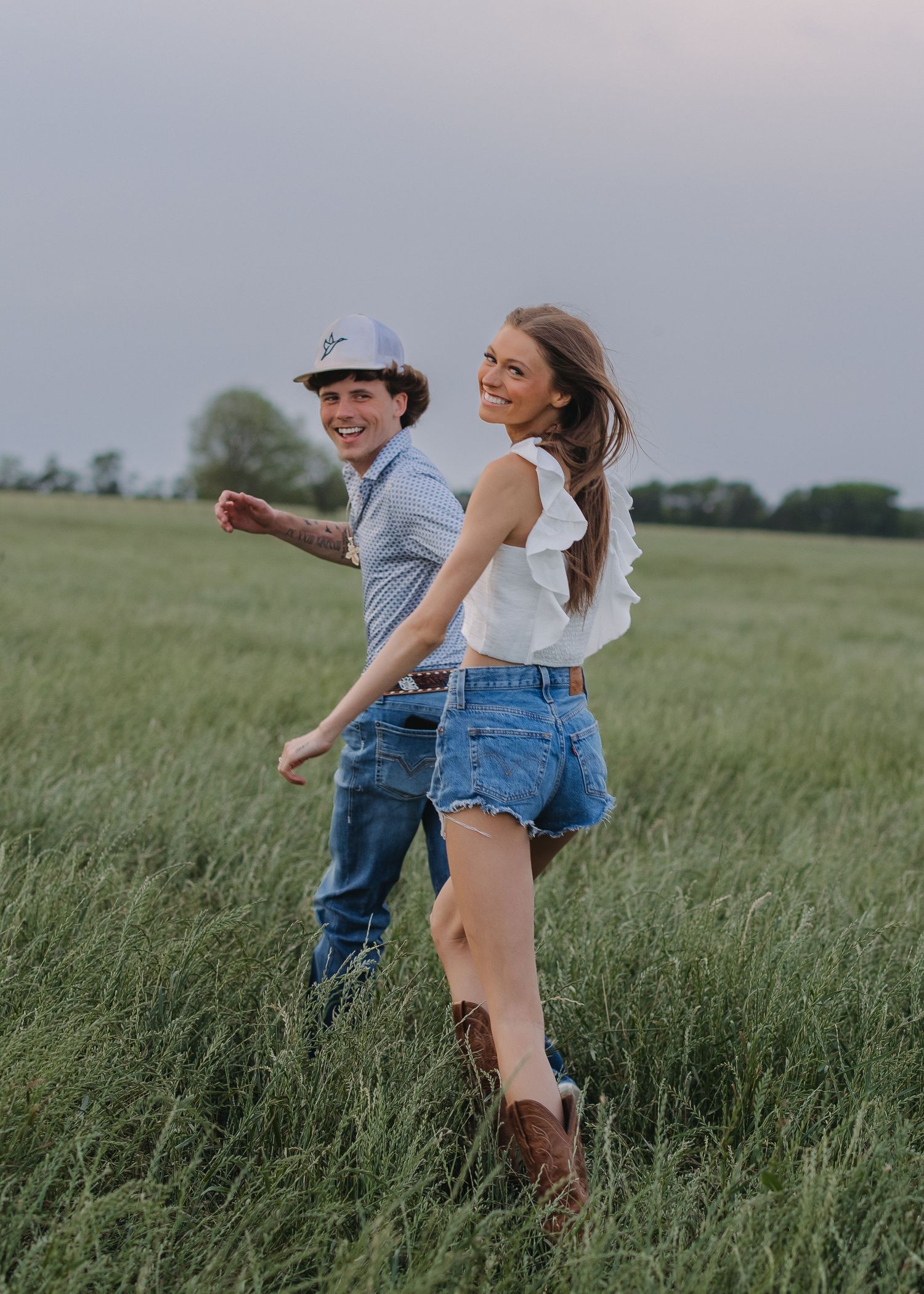 Happy couple walking through green field, woman in denim shorts and white top with cowboy boots.