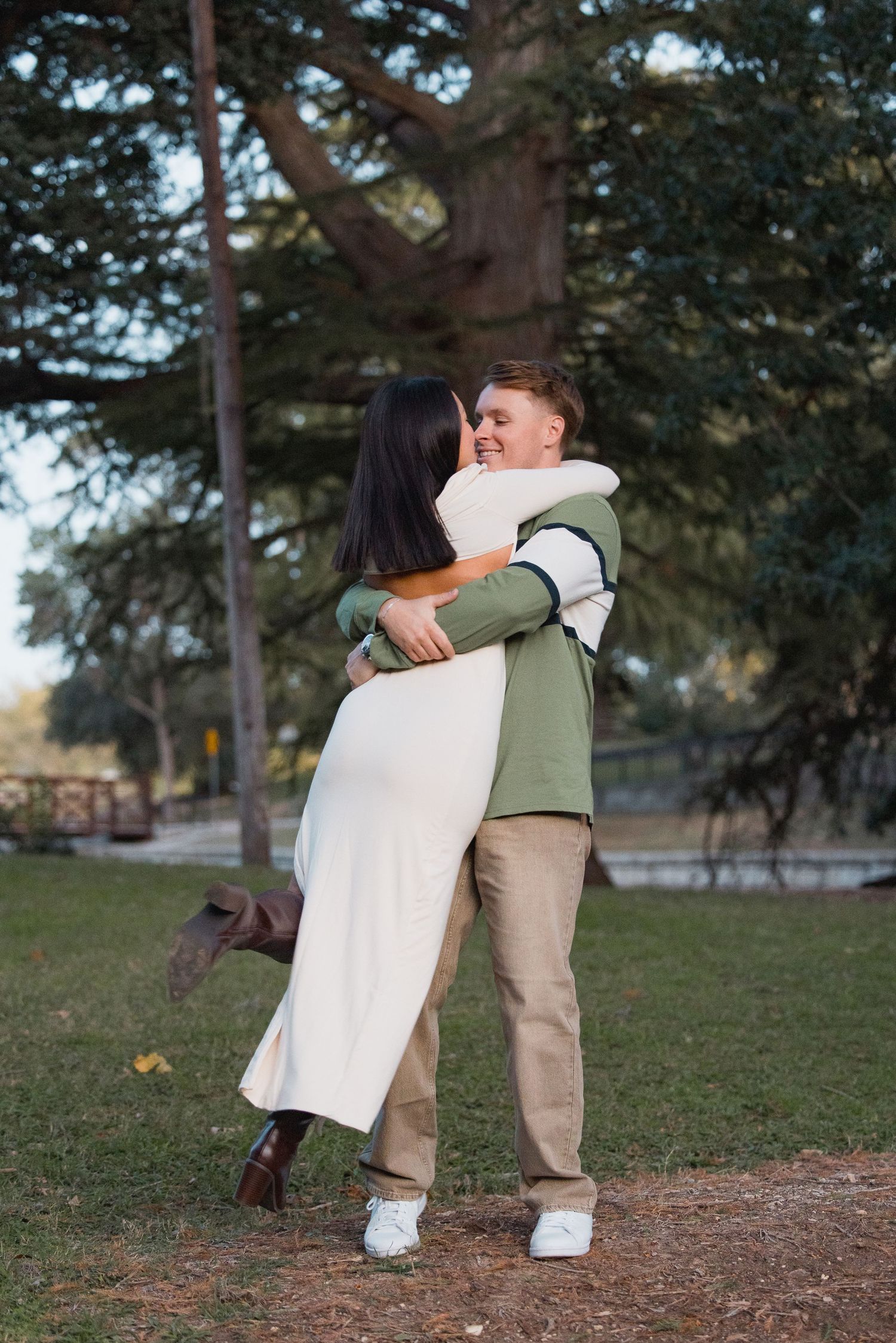 Couple sharing a joyful spinning hug in a sunlit park with tall trees in the background.
