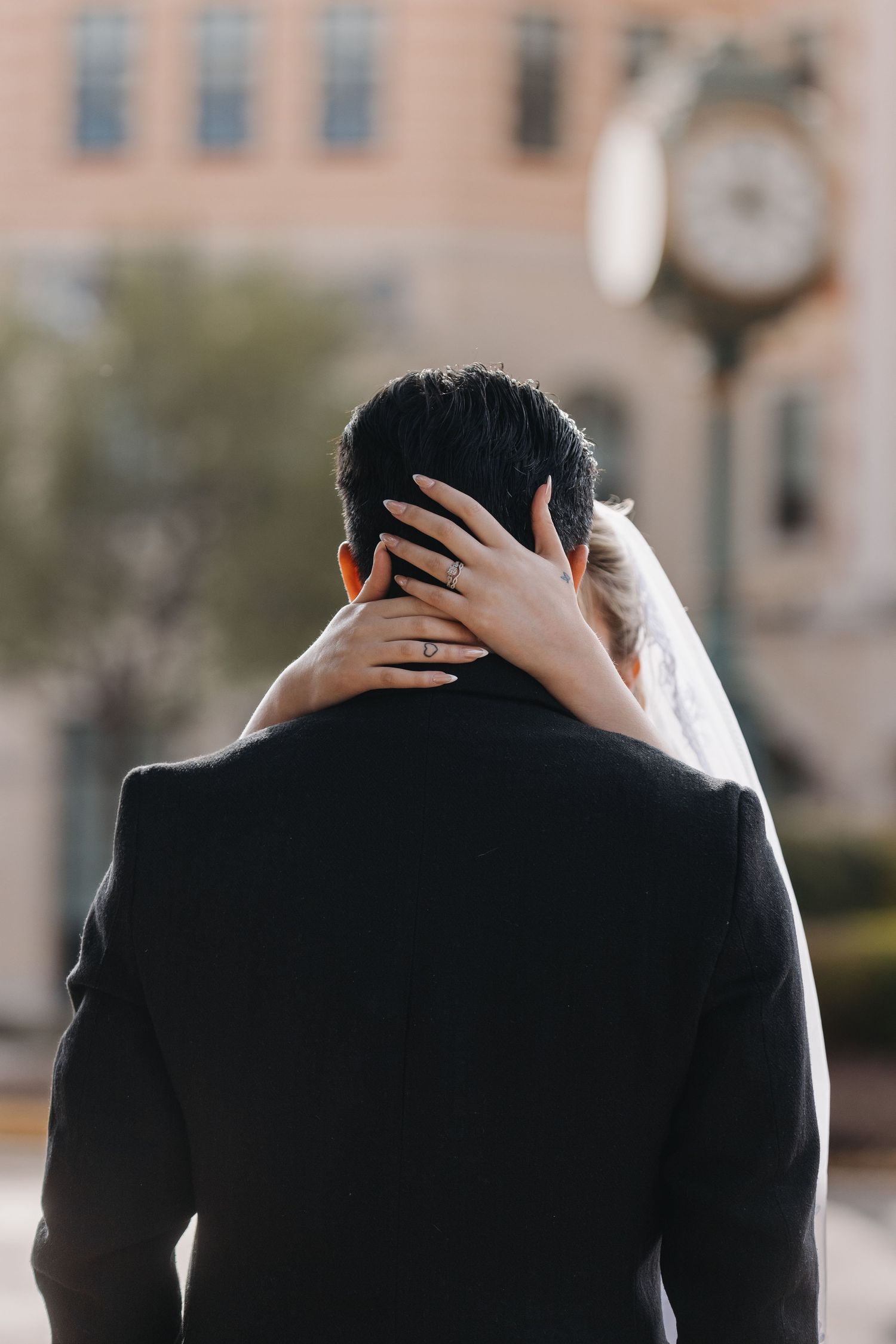 Bride with lace veil places hand on grooms neck as they stand before a clock tower outdoors.