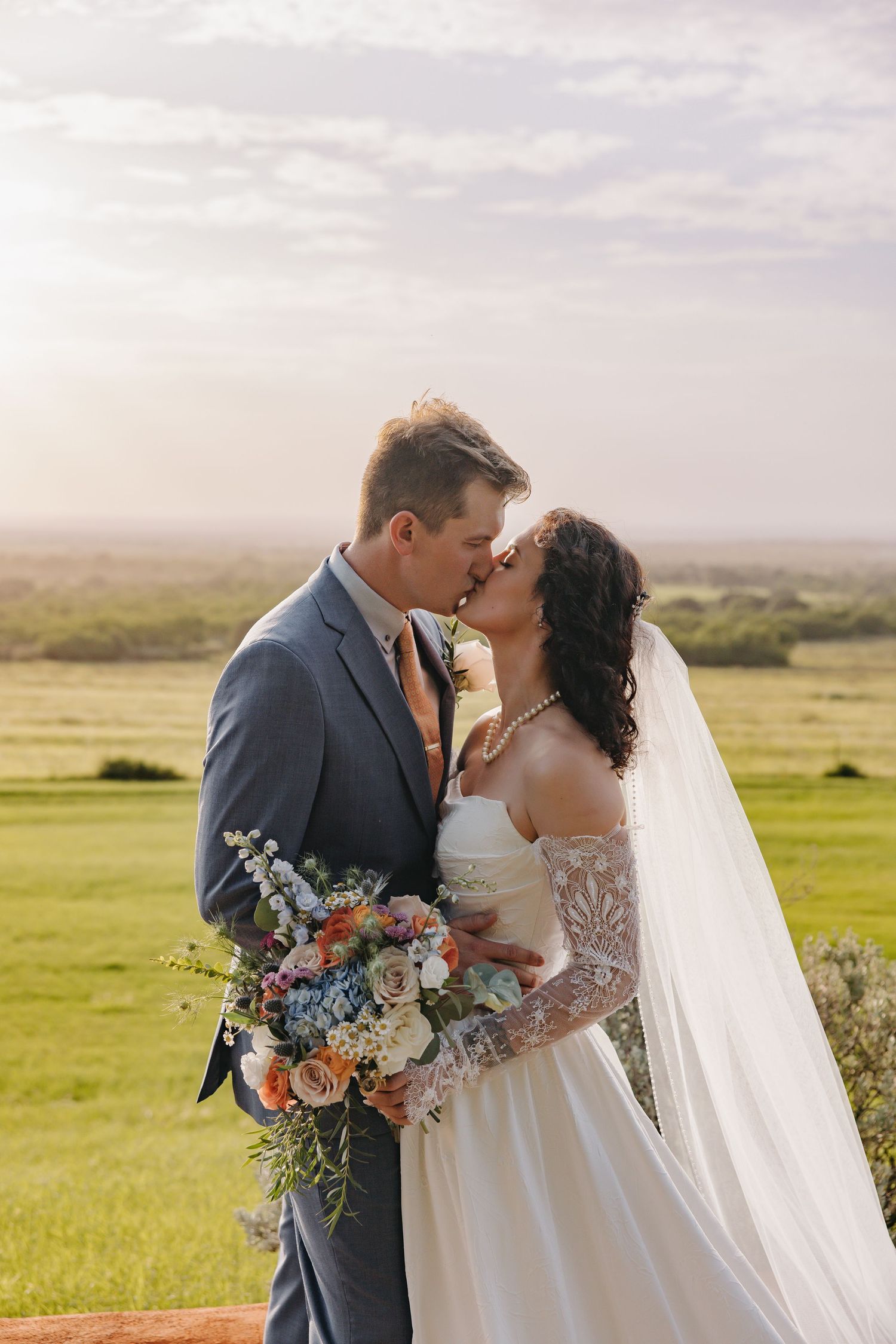Bride and groom kissing outdoors holding colorful floral bouquet at sunset.