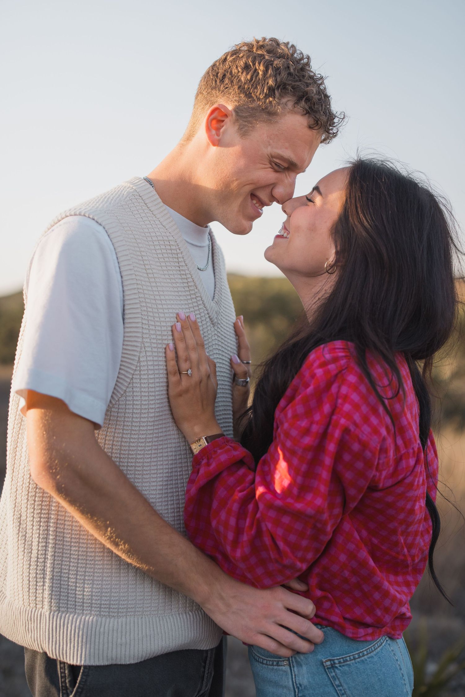 Smiling couple touching foreheads in golden sunlight, woman in pink plaid shirt and man in cream vest.