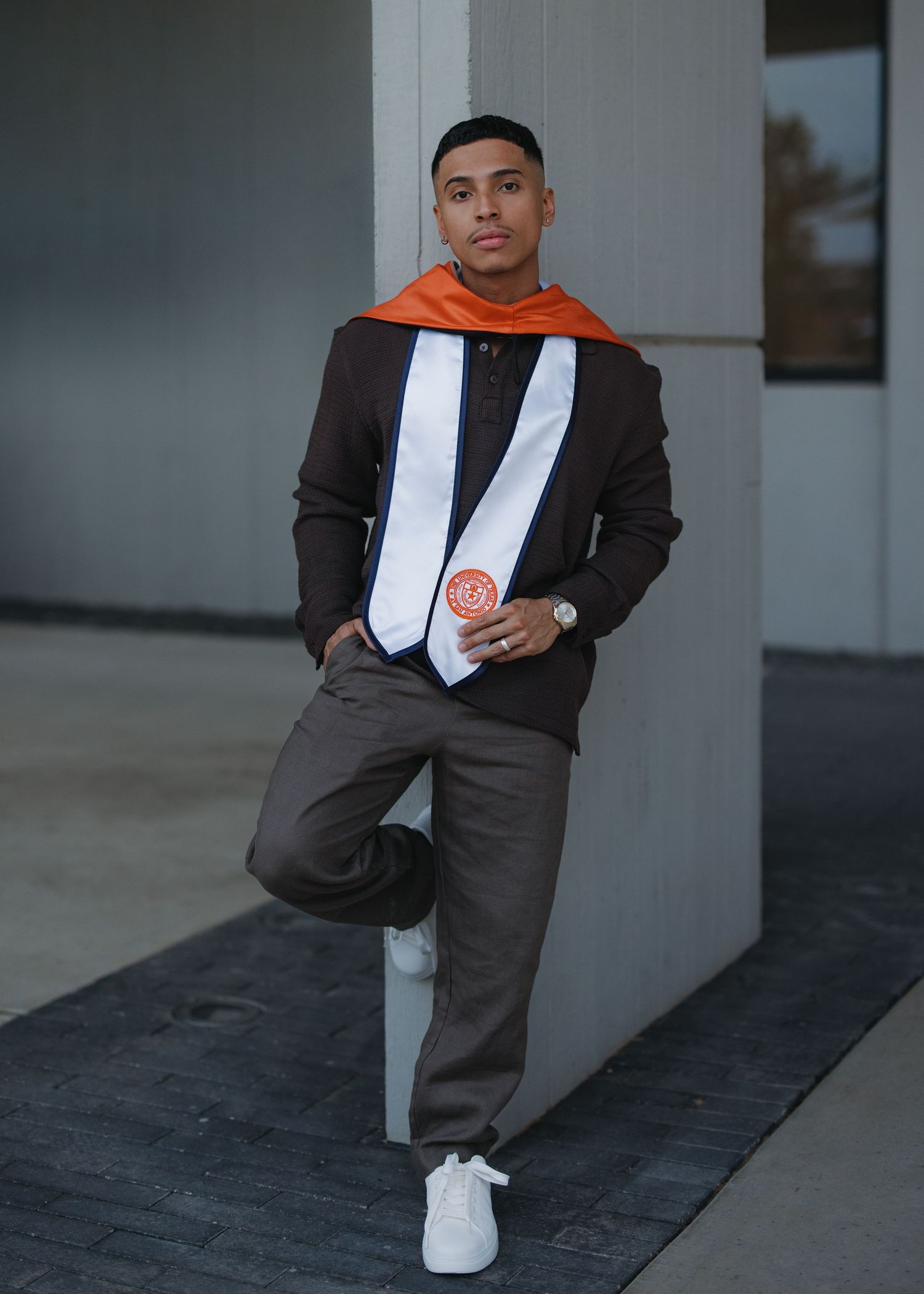 Young man in graduation stole and blazer posing confidently against concrete pillar outdoors.