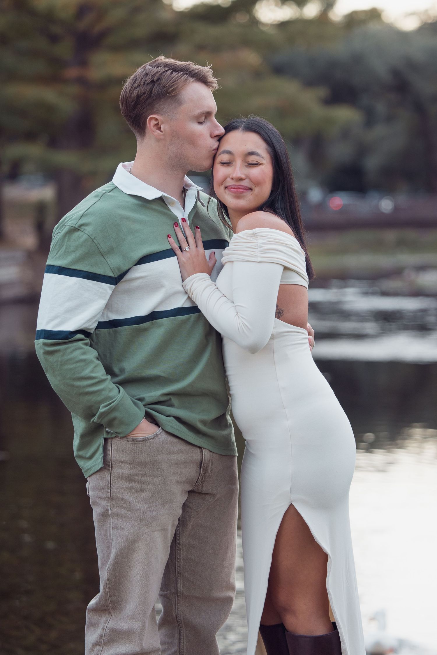 Man in green polo kissing woman on cheek as she smiles contentedly by a tranquil park pond.
