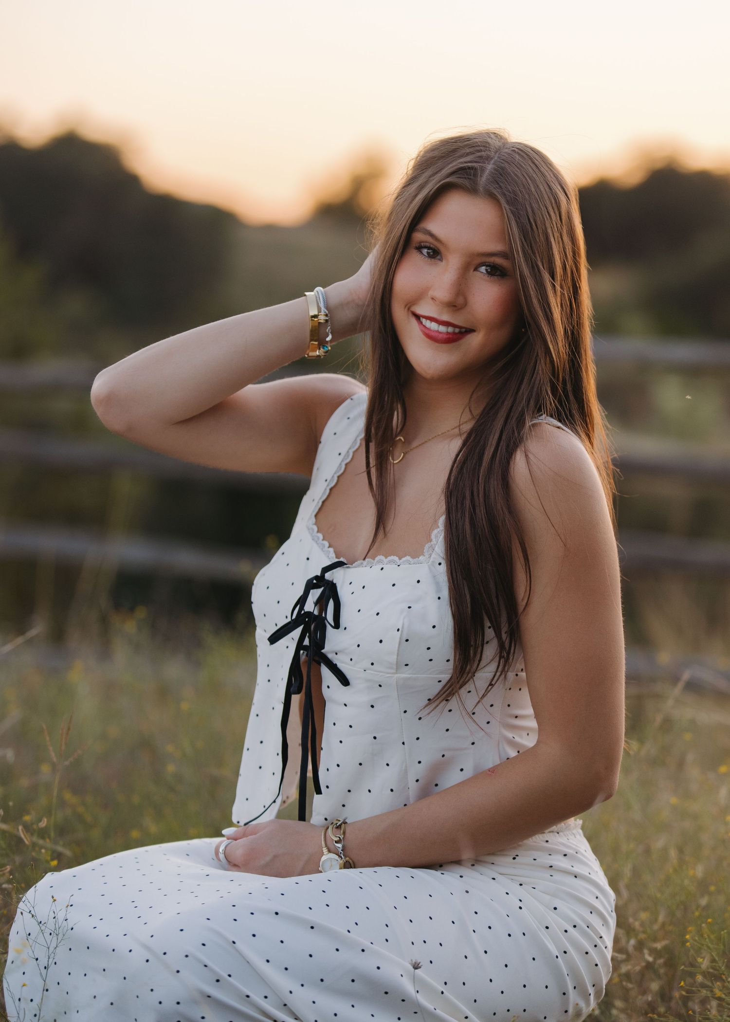 Smiling brunette teen in white polka dot sundress poses at golden sunset near rustic wooden fence outdoors.