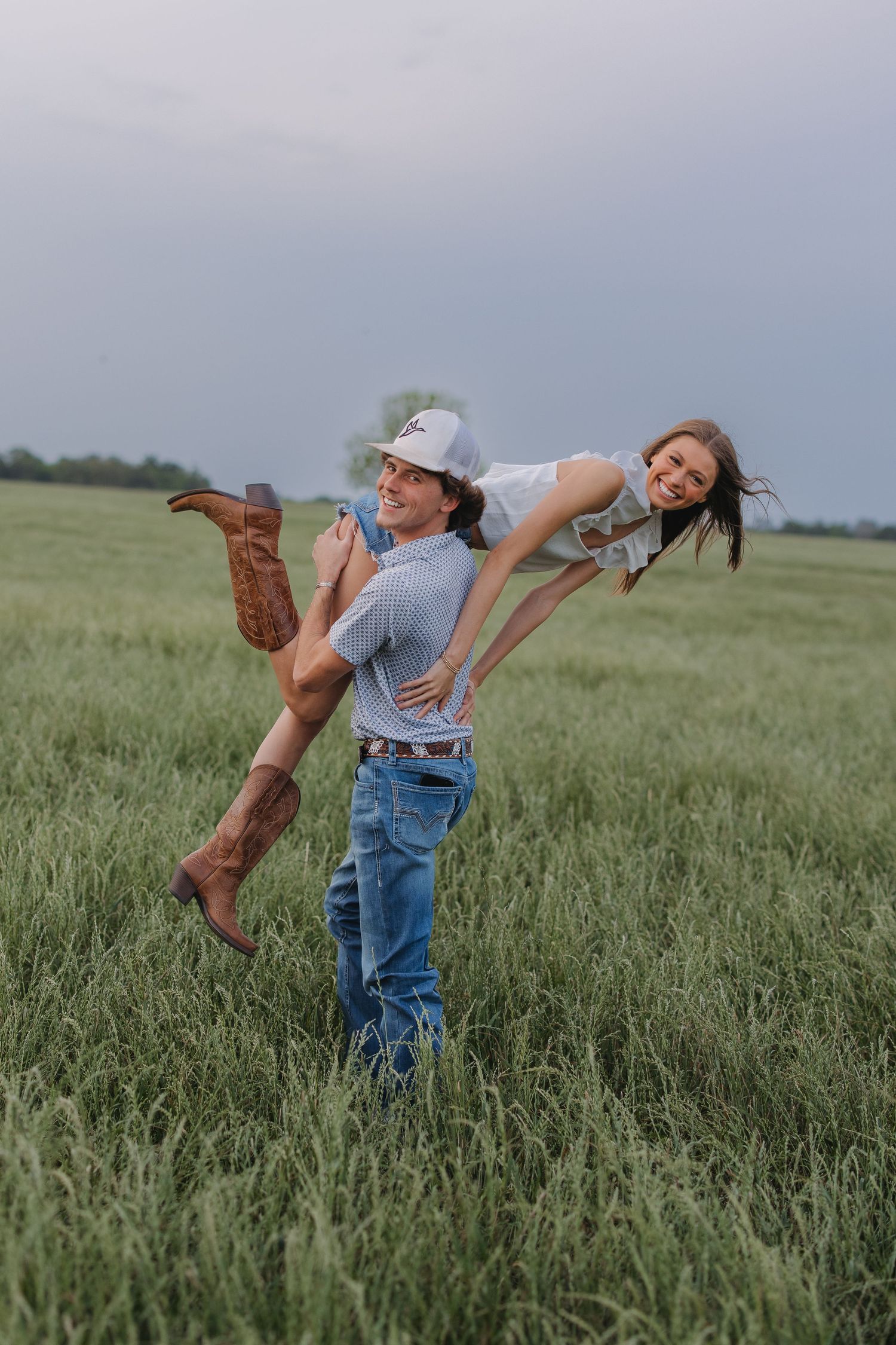 Man joyfully lifting laughing woman in cowboy boots over his shoulder in a green field.