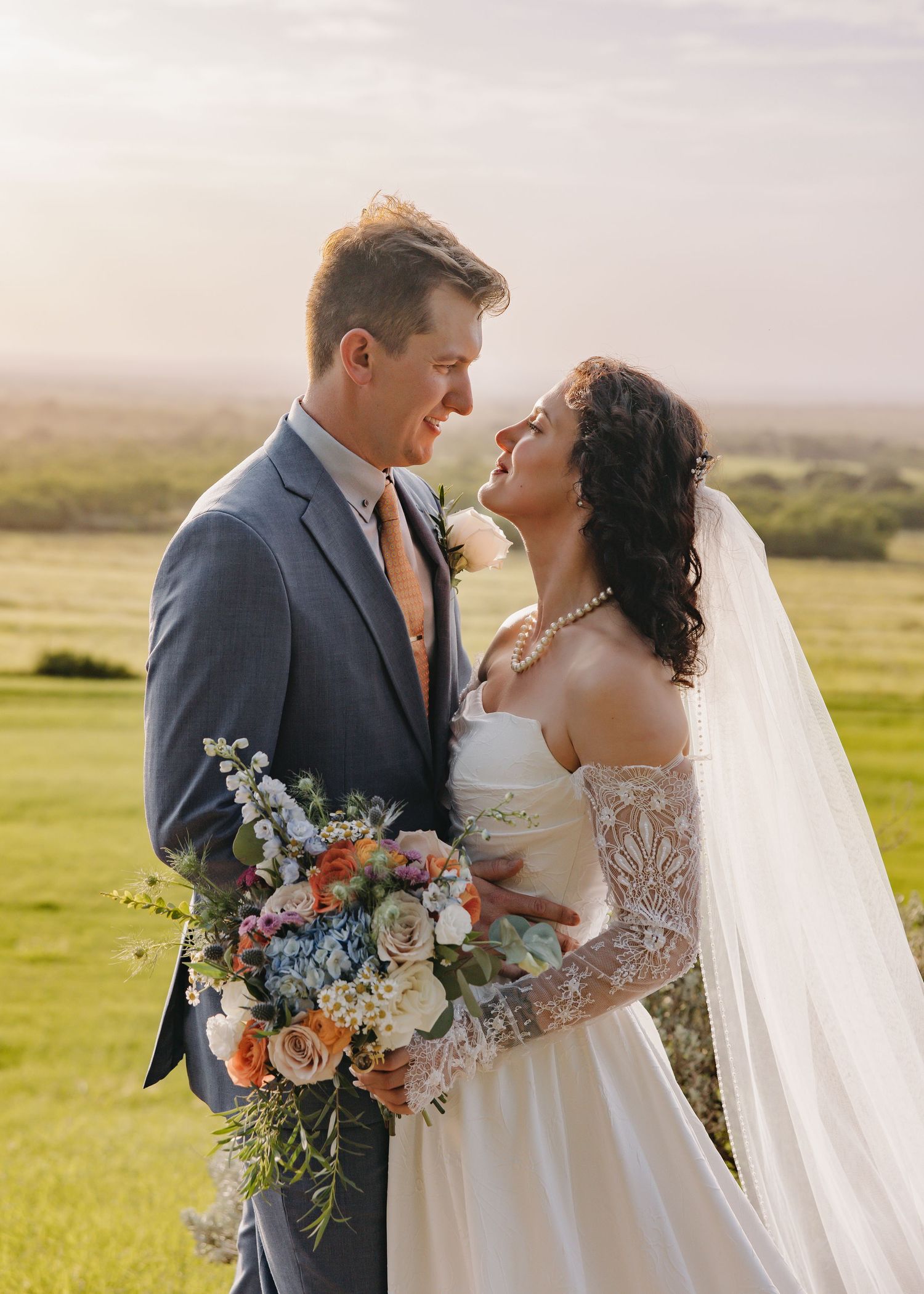 Bride and groom sharing romantic moment at golden hour in lush green field with colorful bouquet.