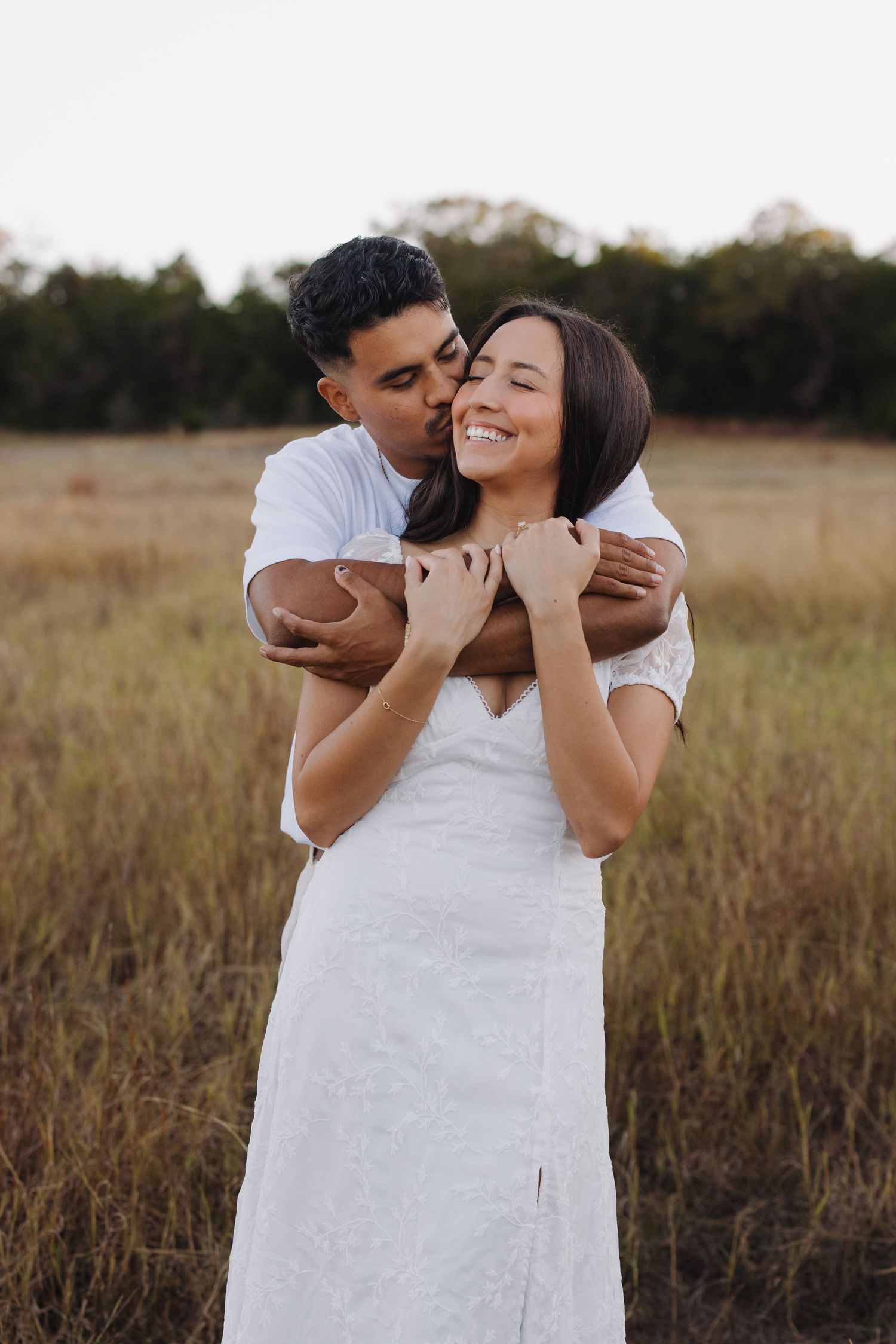 Couple embracing in golden field, man kissing woman's cheek as she smiles, both wearing white.