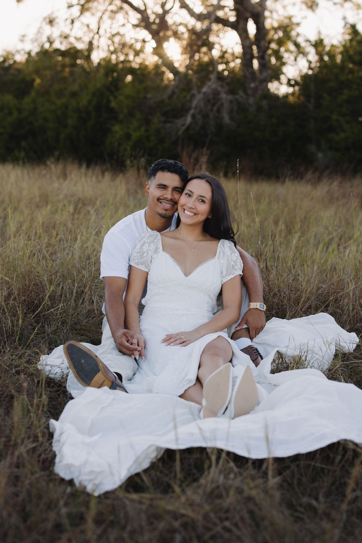 Couple in white outfits sitting on a blanket in a golden grass field at sunset, smiling warmly at the camera.